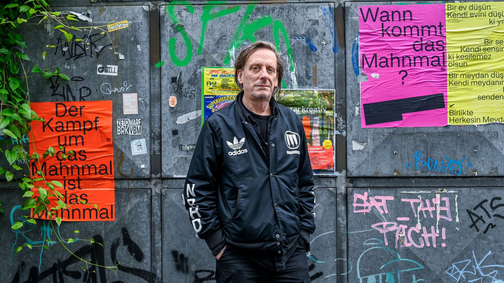 Ein Mann in schwarzer Adidas-Jacke steht vor einem Metalltor, auf dem Plakate kleben, die das fehlende Mahnmal zum Thema haben.