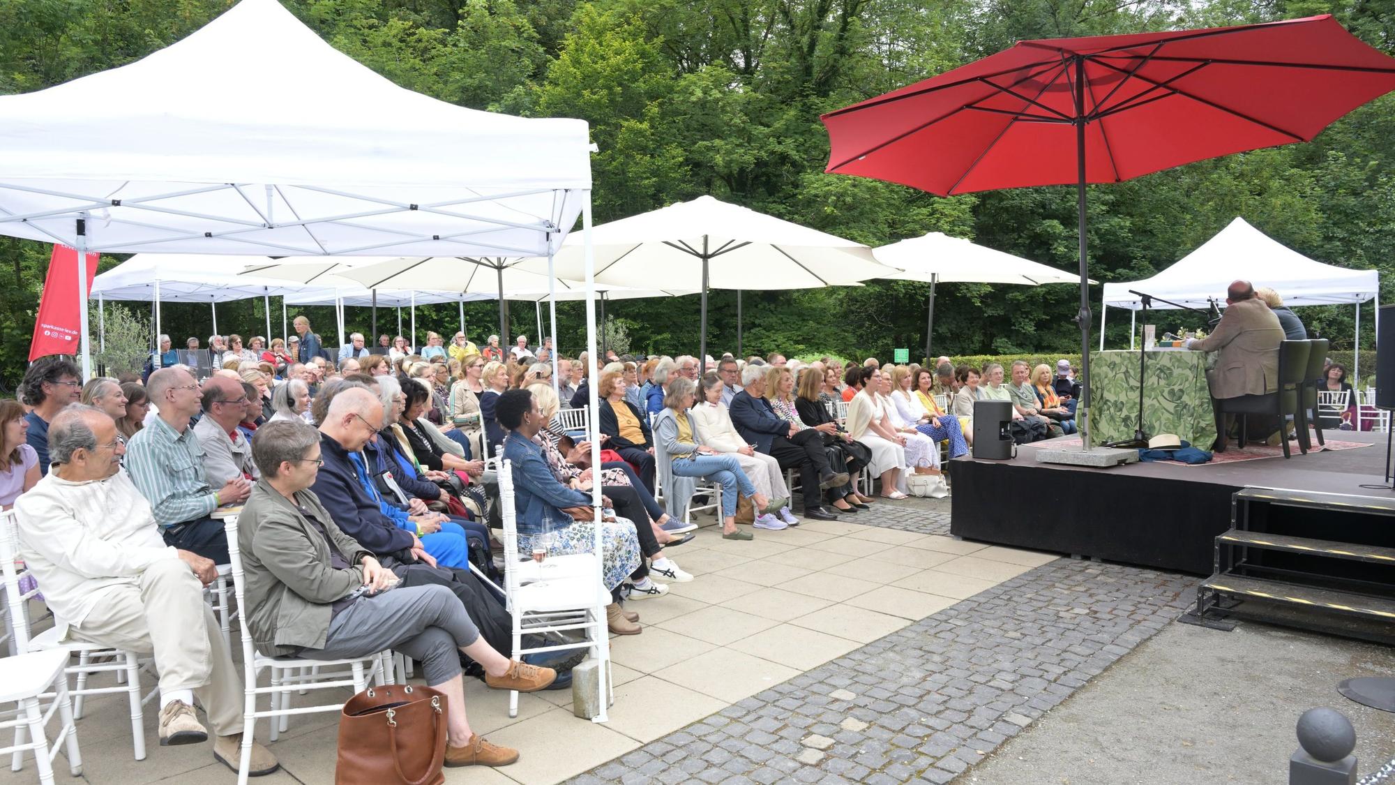 Eine Open-Air-Bühne mit weißen und roten Sonnenschirmen und Publikum auf weißen Stühlen.