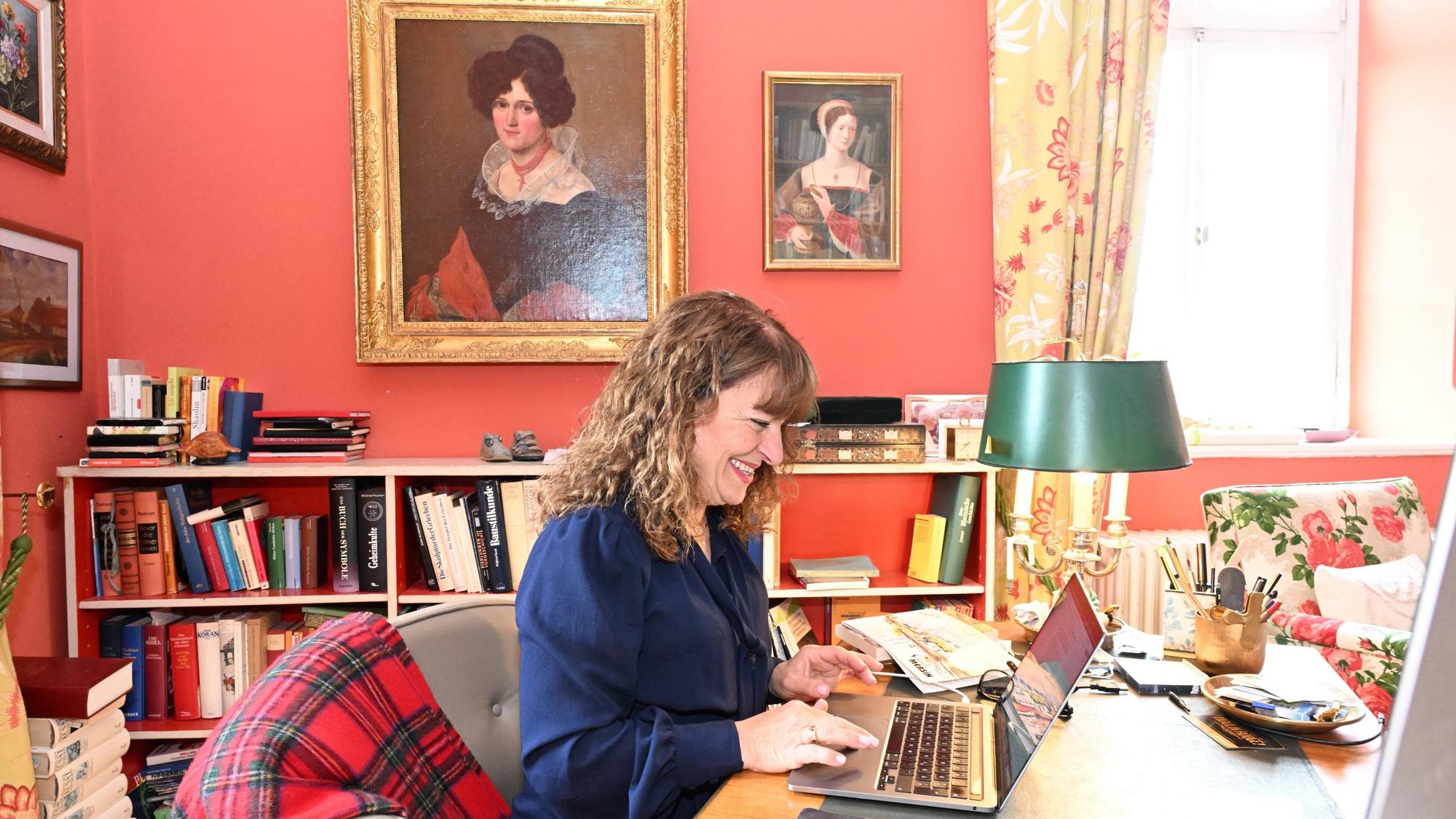 Eine Frau sitzt an einem Laptop in einem Arbeitszimmer mit historischen Ölporträts an den terrakottafarbenen Wänden.