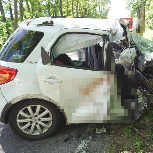 Ein weißes Auto steht völlig zerstört am Straßenrand vor einem Baum.