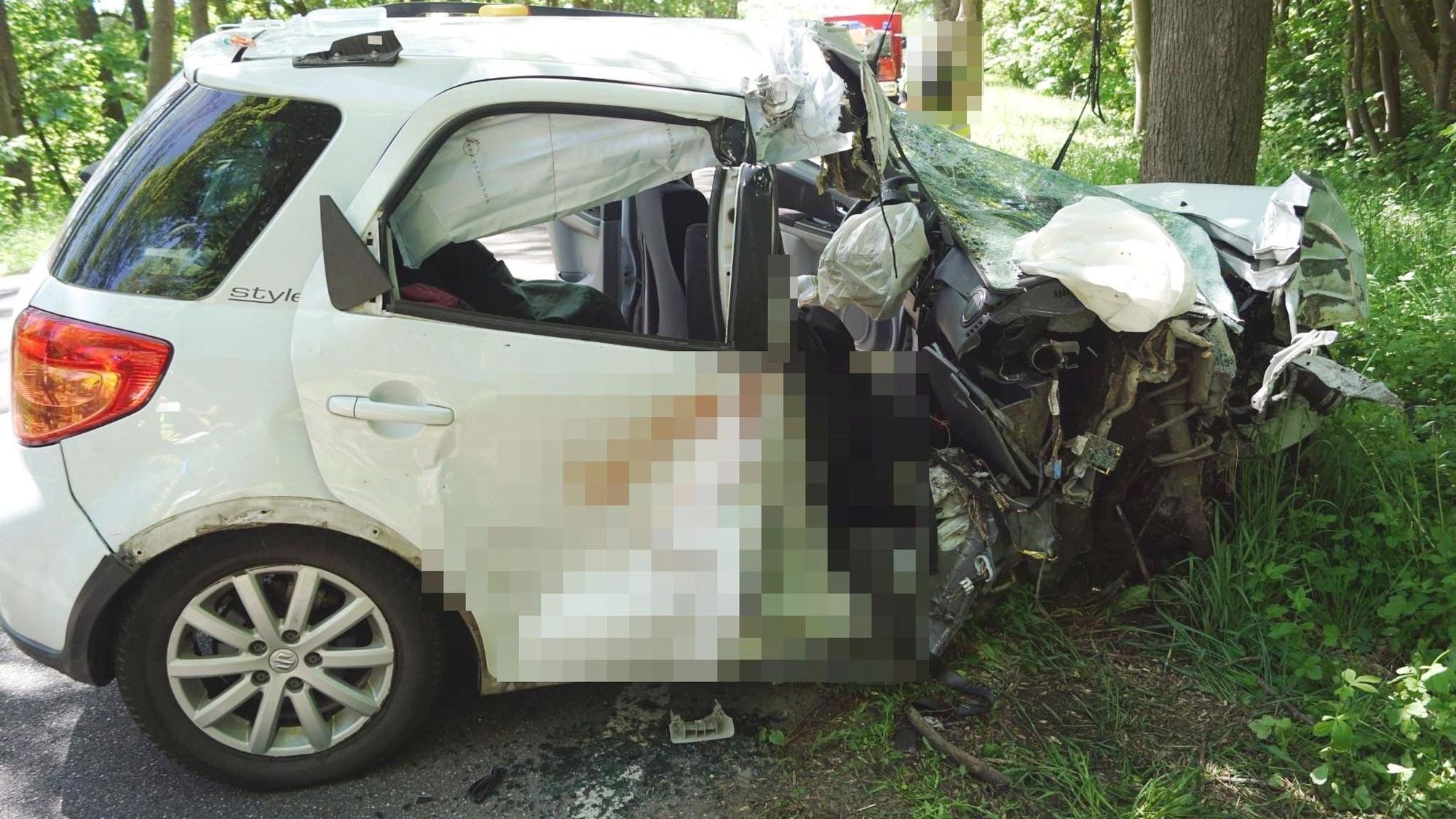 Ein weißes Auto steht völlig zerstört am Straßenrand vor einem Baum.