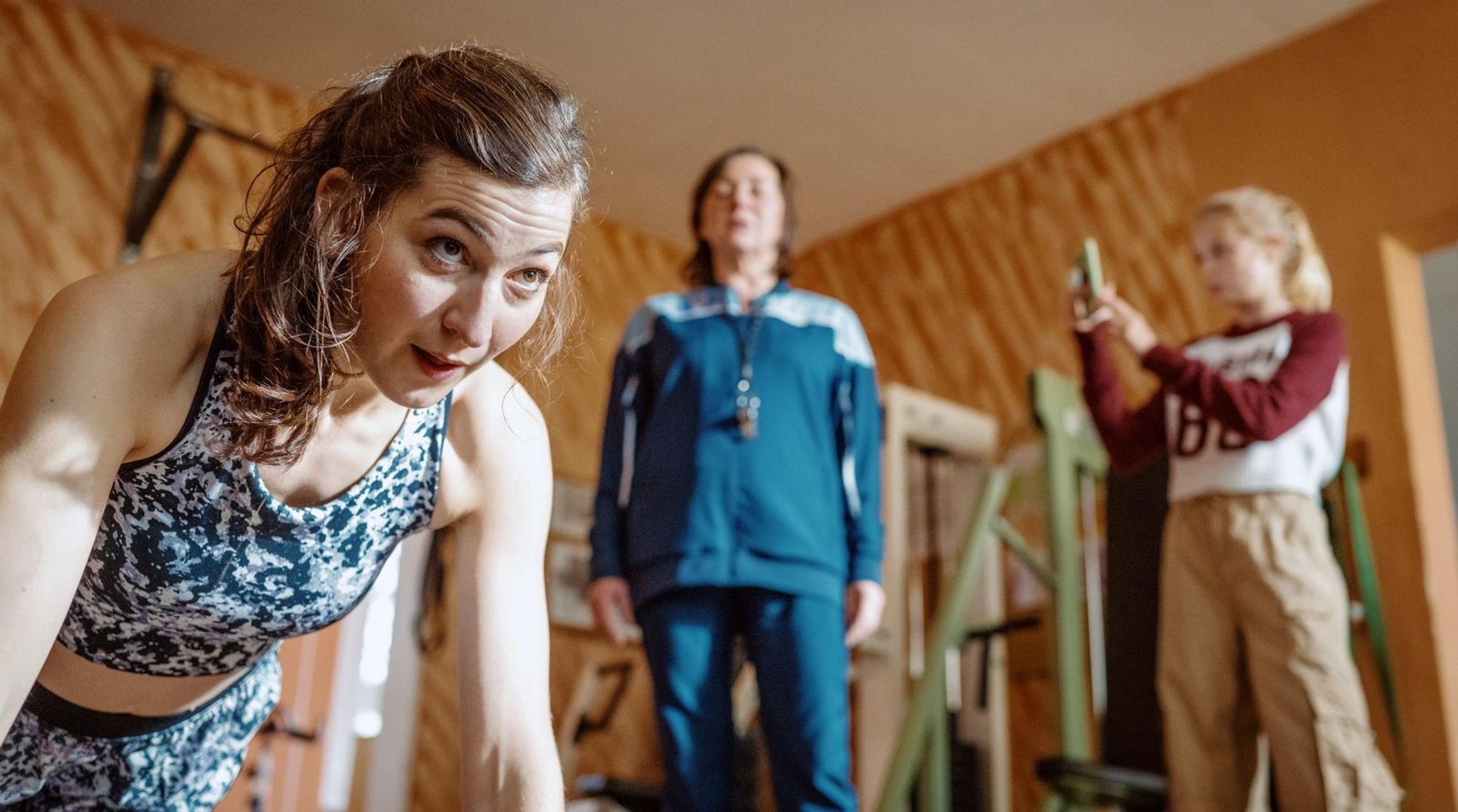 „Fit in Gerlau“ soll über Social Media beworben werden: Enkelin Emma (Selma Suckfüll, rechts) filmt Mitarbeiterin Katleen (Ann-Sophie Schindler, links). Oma Christa (Steffi Kühnert) gibt die Einpeitscherin. (Bild: © ARD Degeto Film/Adrian Gross)