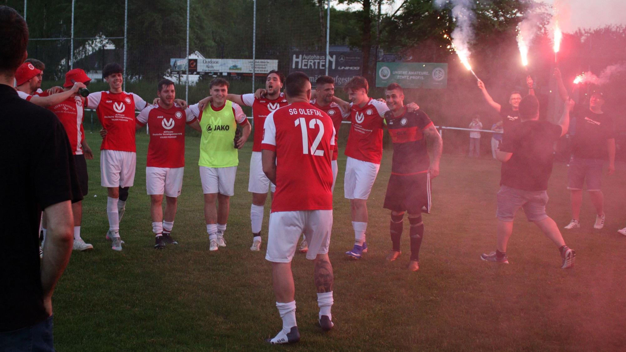 Spieler und Fans der SG Oleftal feiern den Wiederaufsteig. Im Vordergrund steht Sascha Haas mit dem Rücken zum Fotografen, in der Hand hält er eine Sektflasche. Am rechten Bildrand stehen Mitglieder der "Westkurve" mit roten Leuchtfackeln.