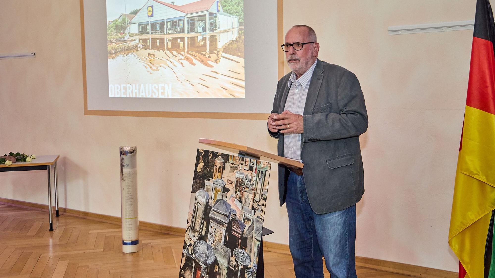 Klaus Jansen steht an einem Rednerpult, links von ihm steht die Flutkerze und an der Wand dahinter wird eine Präsentation gezeigt.