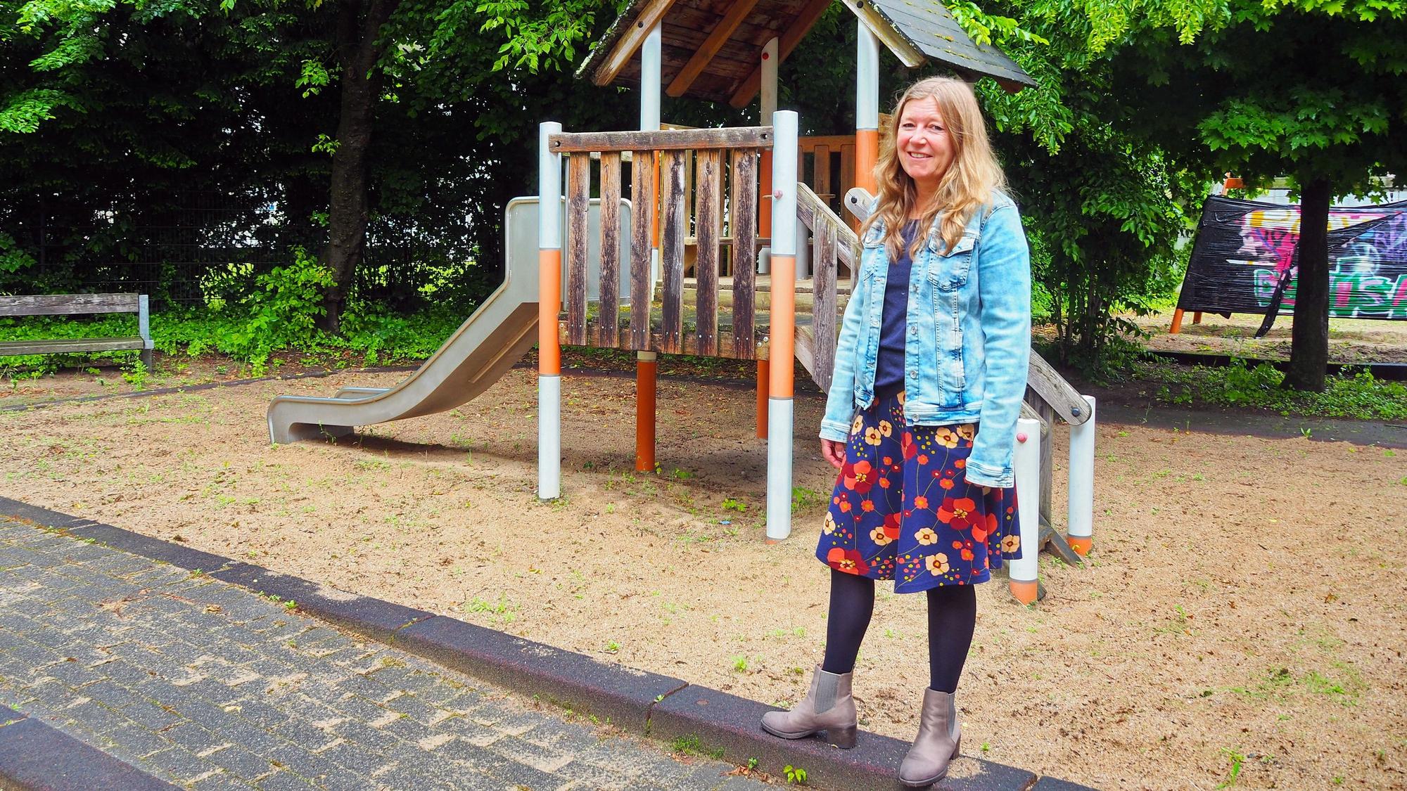 Eine Frau in Jeansjacke und Blumenkleid steht vor einem Spielehaus auf einer Sandfläche eines Spielplatzes.