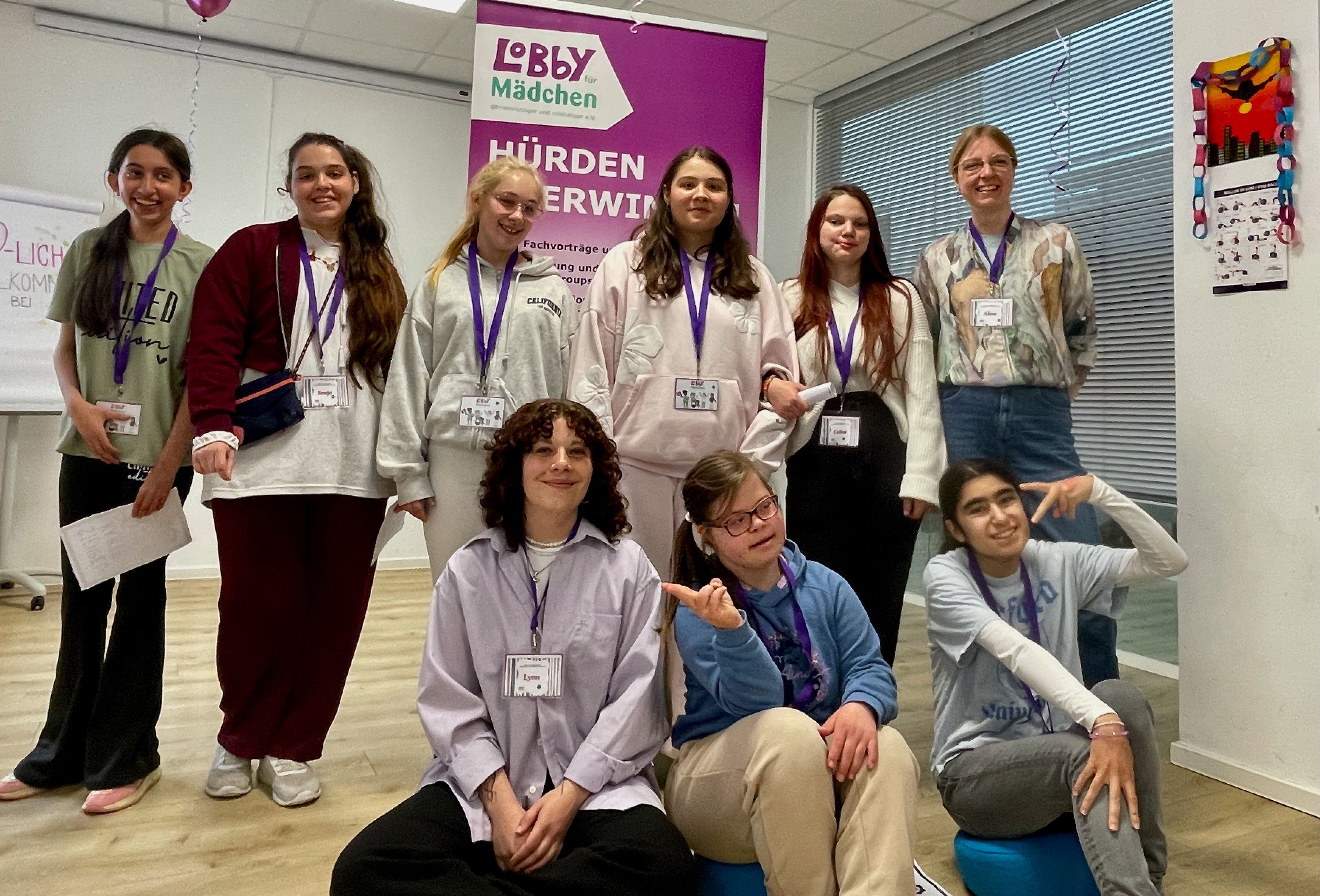 Junge Frauen stehen und sitzen vor einem Aufsteller, auf dem steht „Lobby für Mädchen Hürden überwinden“.