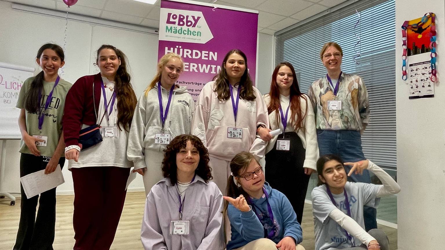 Junge Frauen stehen und sitzen vor einem Aufsteller, auf dem steht „Lobby für Mädchen Hürden überwinden“.