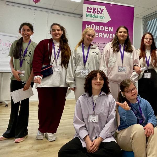Junge Frauen stehen und sitzen vor einem Aufsteller, auf dem steht „Lobby für Mädchen Hürden überwinden“.