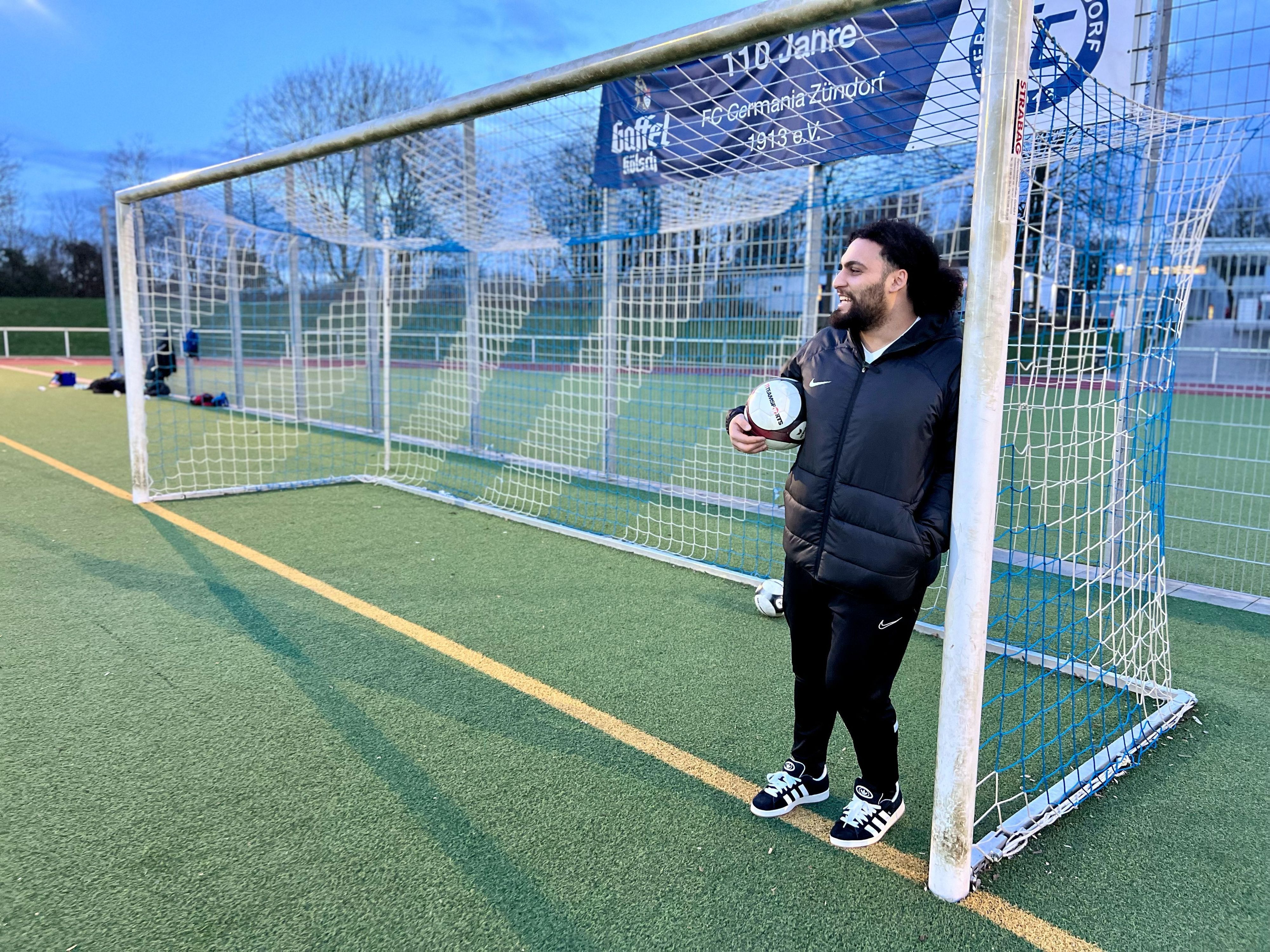 Ein Mann mit dunklem Haar und Trainingsklamotten steht mit Fußball unter dem Arm an einen Pfosten eines Fußballtors gelehnt.