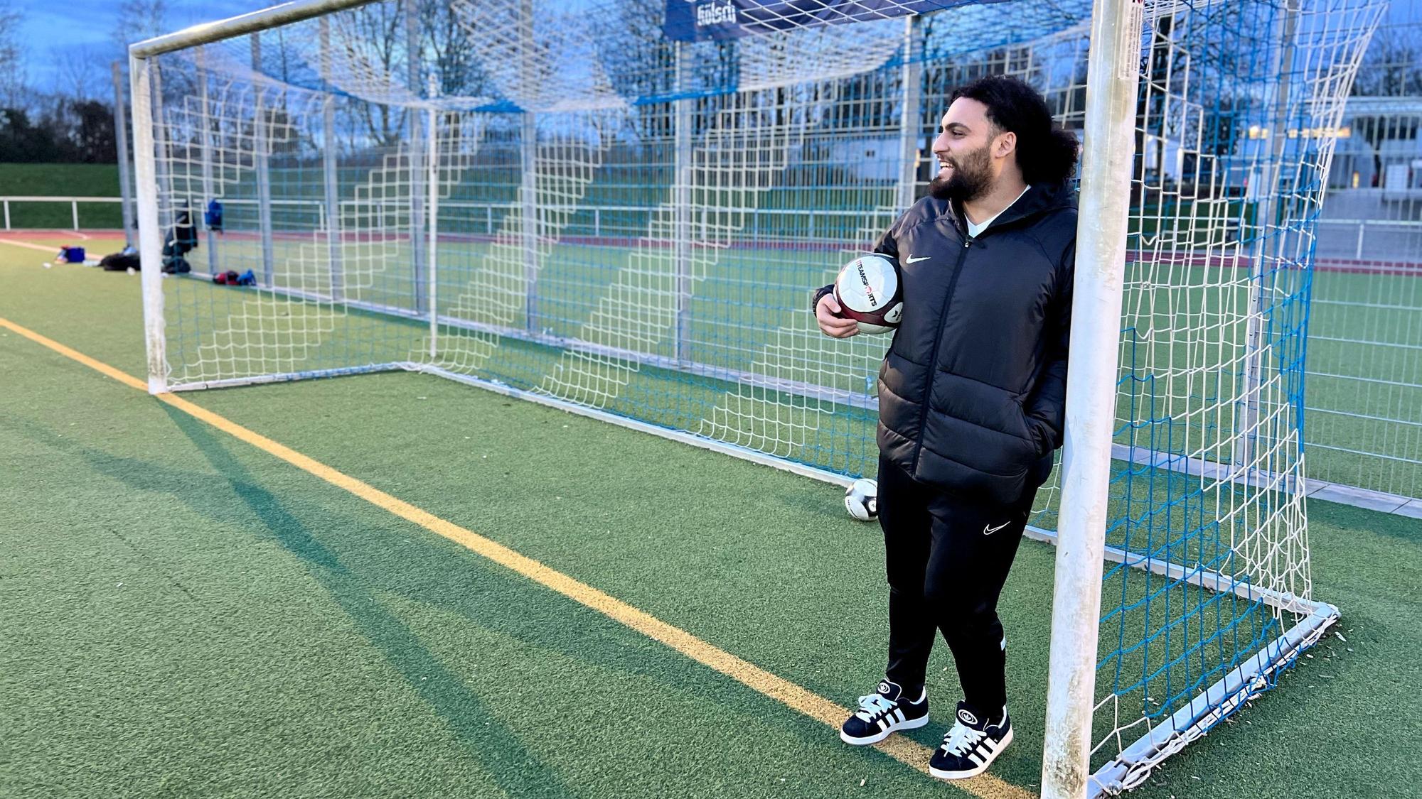 Ein Mann mit dunklem Haar und Trainingsklamotten steht mit Fußball unter dem Arm an einen Pfosten eines Fußballtors gelehnt.