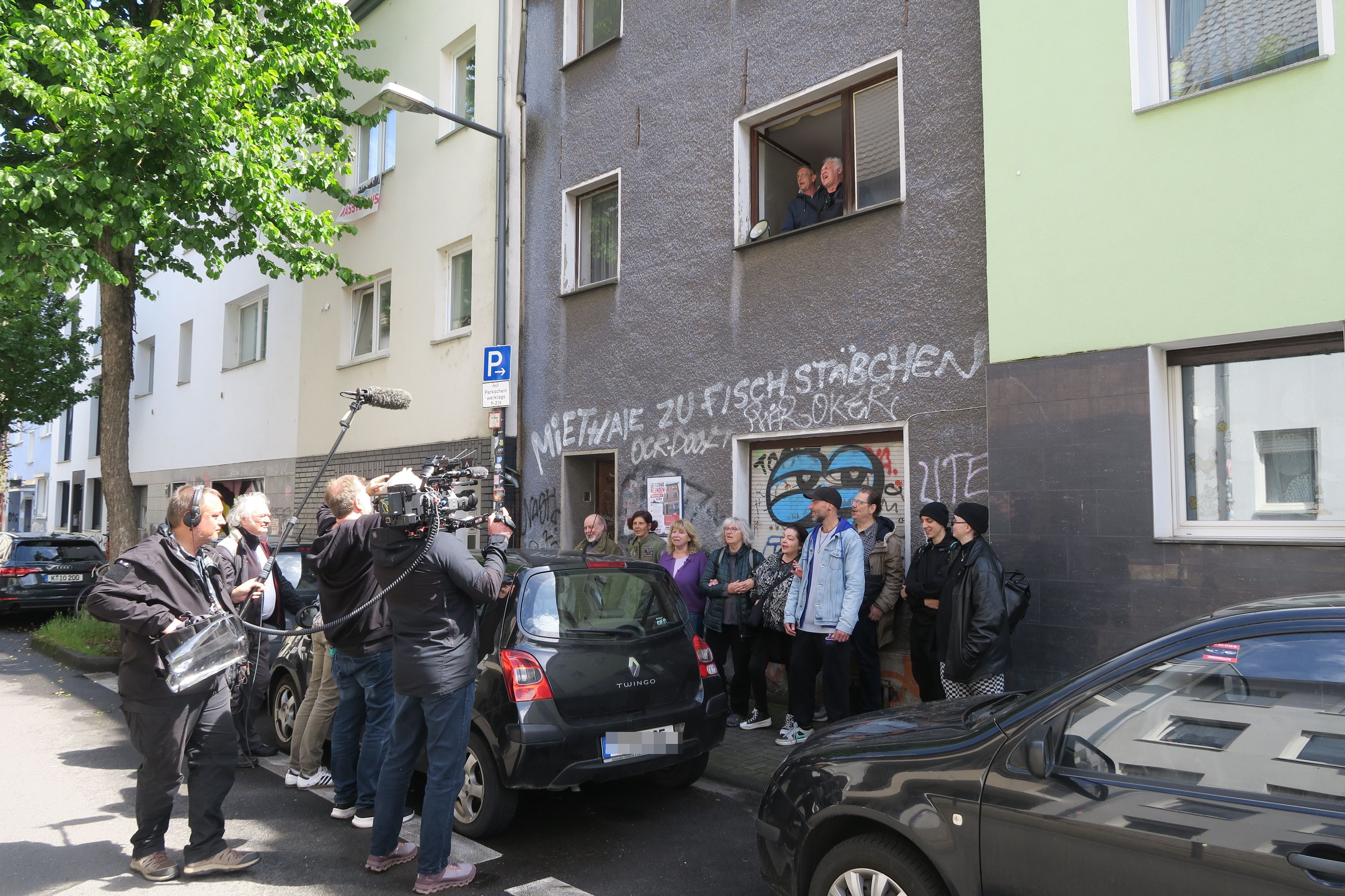Ein Kamerateam filmt eine Menschengruppe, die sich vor einem Haus versammelt hat.