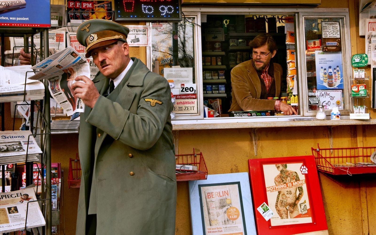 Der erste Weg für den neu erwachten Hitler (Oliver Masucci, links) zu einem Kiosk, an dem ihm seine Situation bewusst wird. Er lebt nun im Jahr 2014. Den „Krämer“ spielt Lars Rudolph. (Bild:  2015 Constantin Film Verleih GmbH)