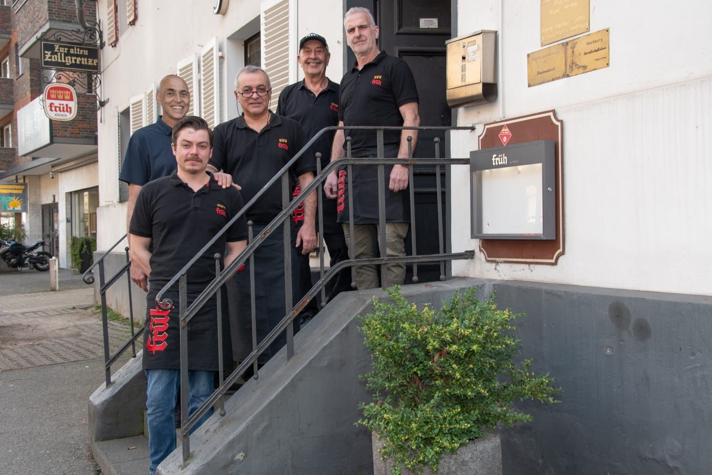 Fünf Männer in schwarzen T-Shirts und Kellnerschürzen stehen auf einer kleinen Treppe zum Eingang des Lokals Alte Zollgrenze.