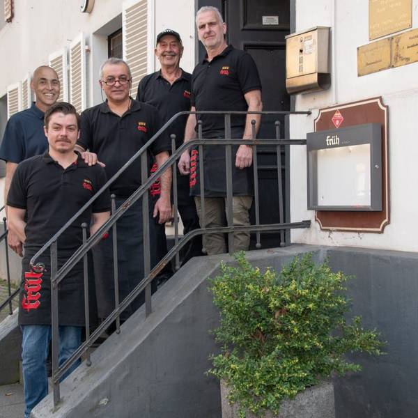 Fünf Männer in schwarzen T-Shirts und Kellnerschürzen stehen auf einer kleinen Treppe zum Eingang des Lokals Alte Zollgrenze.