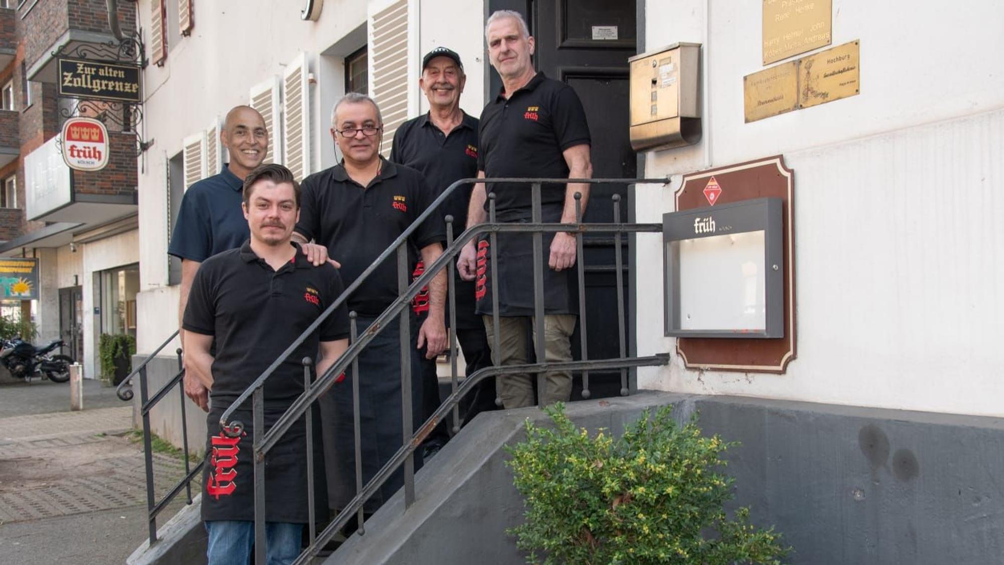 Fünf Männer in schwarzen T-Shirts und Kellnerschürzen stehen auf einer kleinen Treppe zum Eingang des Lokals Alte Zollgrenze.