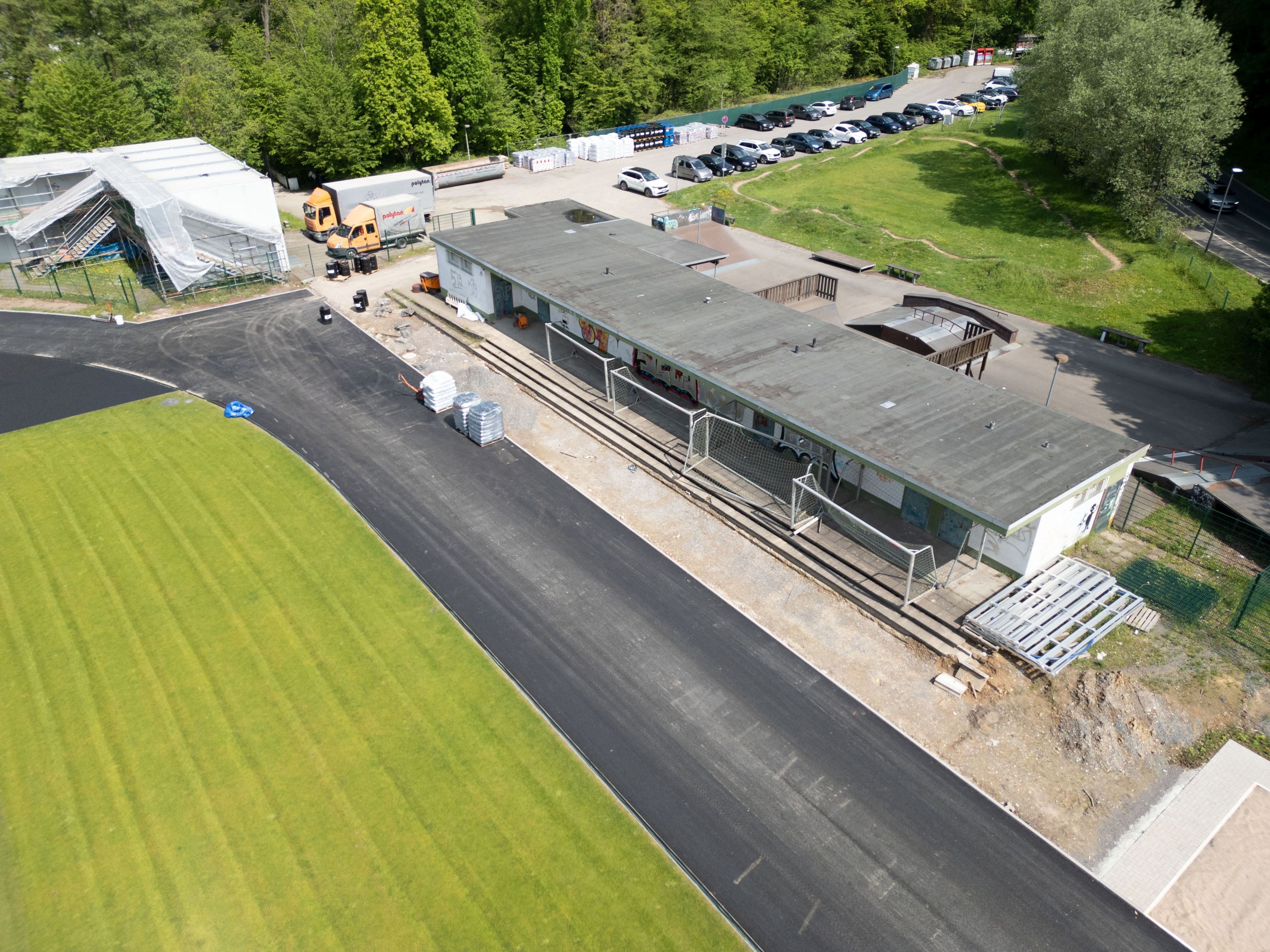 Ein Sportplatz im Umbau. Grüner Rasen ist schon verlegt, die Tartanbahn wird gerade gebaut. Dahinter ein verhülltes Gebäude. Ein Flachbau ist marode.