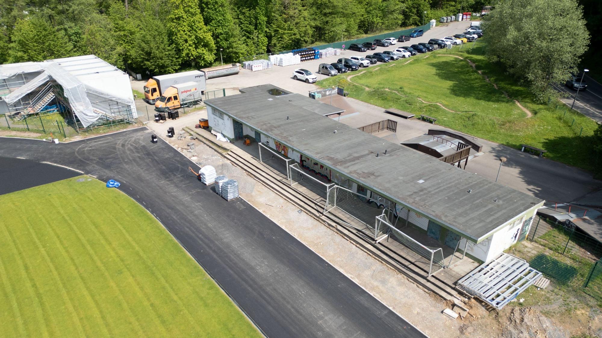 Ein Sportplatz im Umbau. Grüner Rasen ist schon verlegt, die Tartanbahn wird gerade gebaut. Dahinter ein verhülltes Gebäude. Ein Flachbau ist marode.
