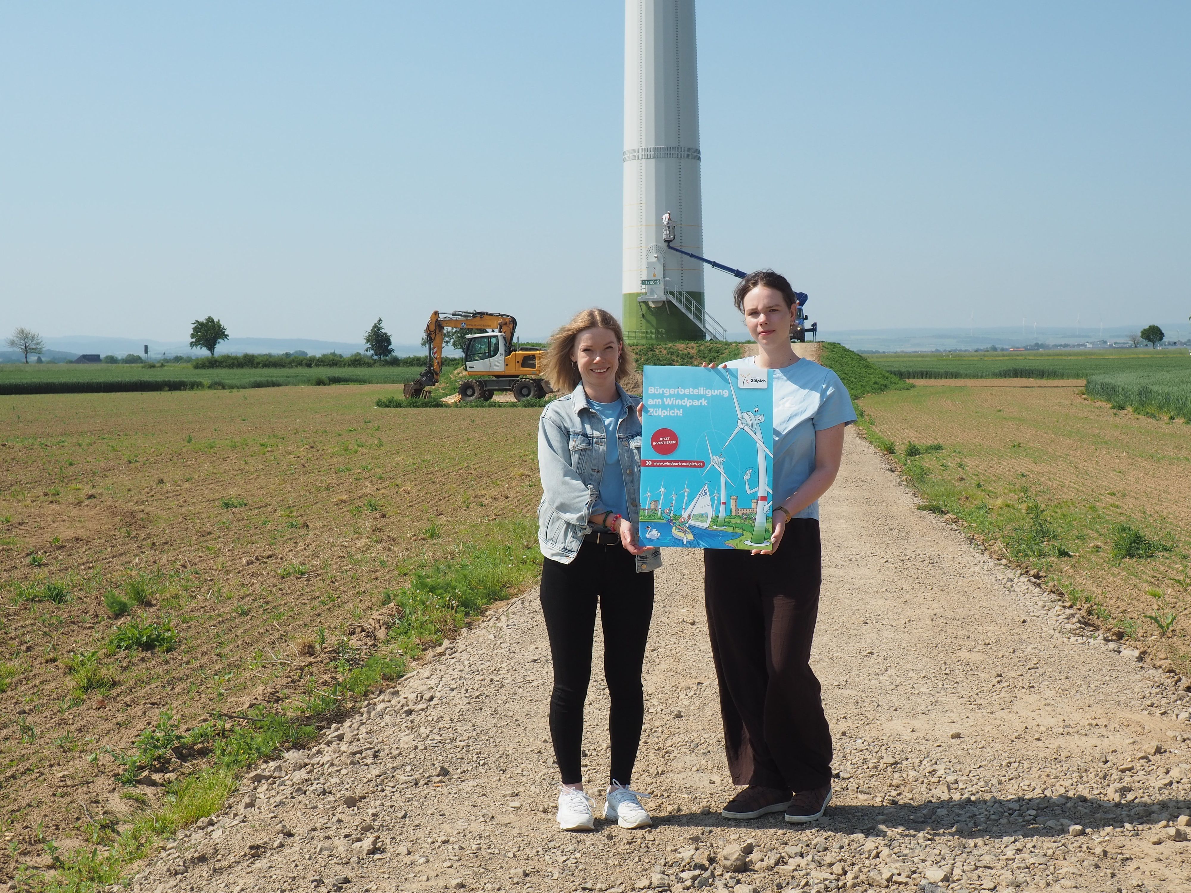 Stefanie Flam und Laura Mertens stehen vor einem Windrad, das gerade angestrichen wird, und halten ein Schild, das für die Bürgerbeteiligung wirbt.