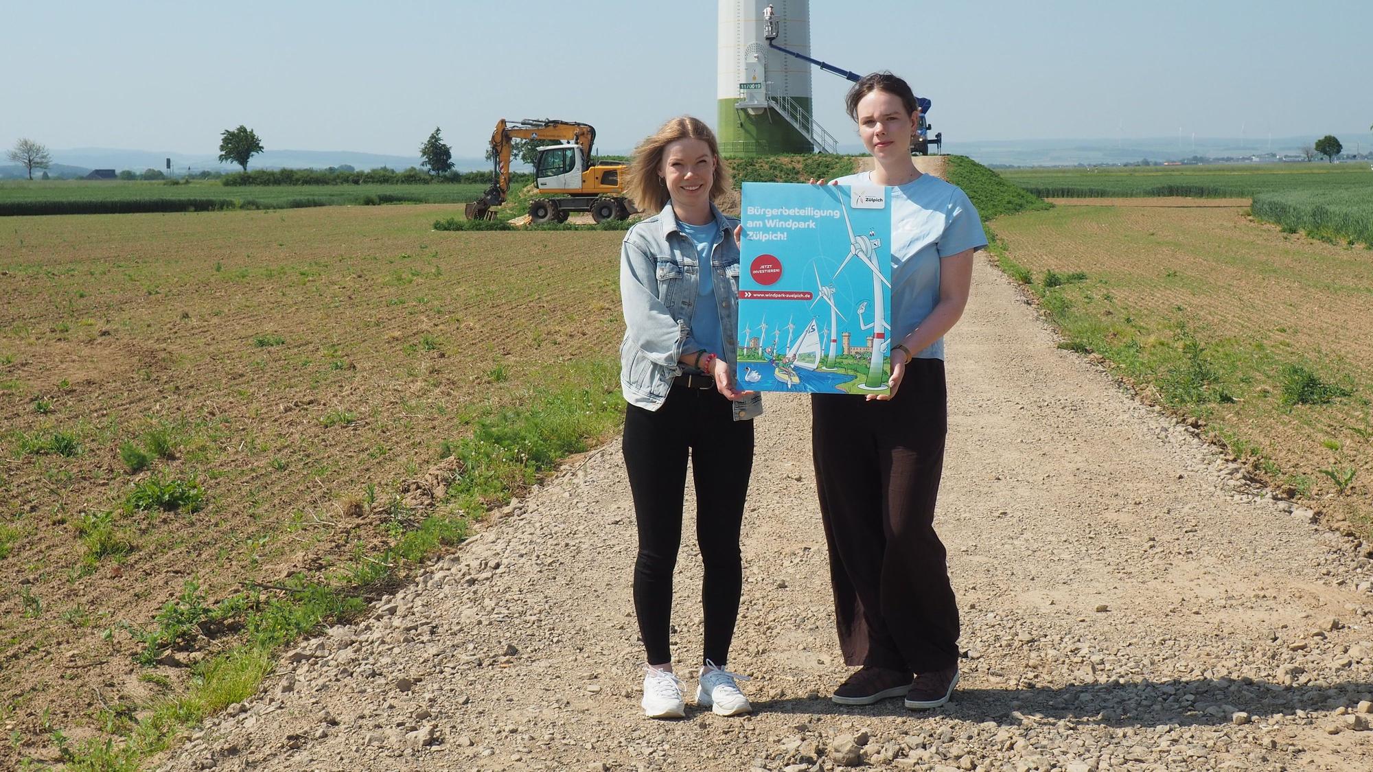 Stefanie Flam und Laura Mertens stehen vor einem Windrad, das gerade angestrichen wird, und halten ein Schild, das für die Bürgerbeteiligung wirbt.
