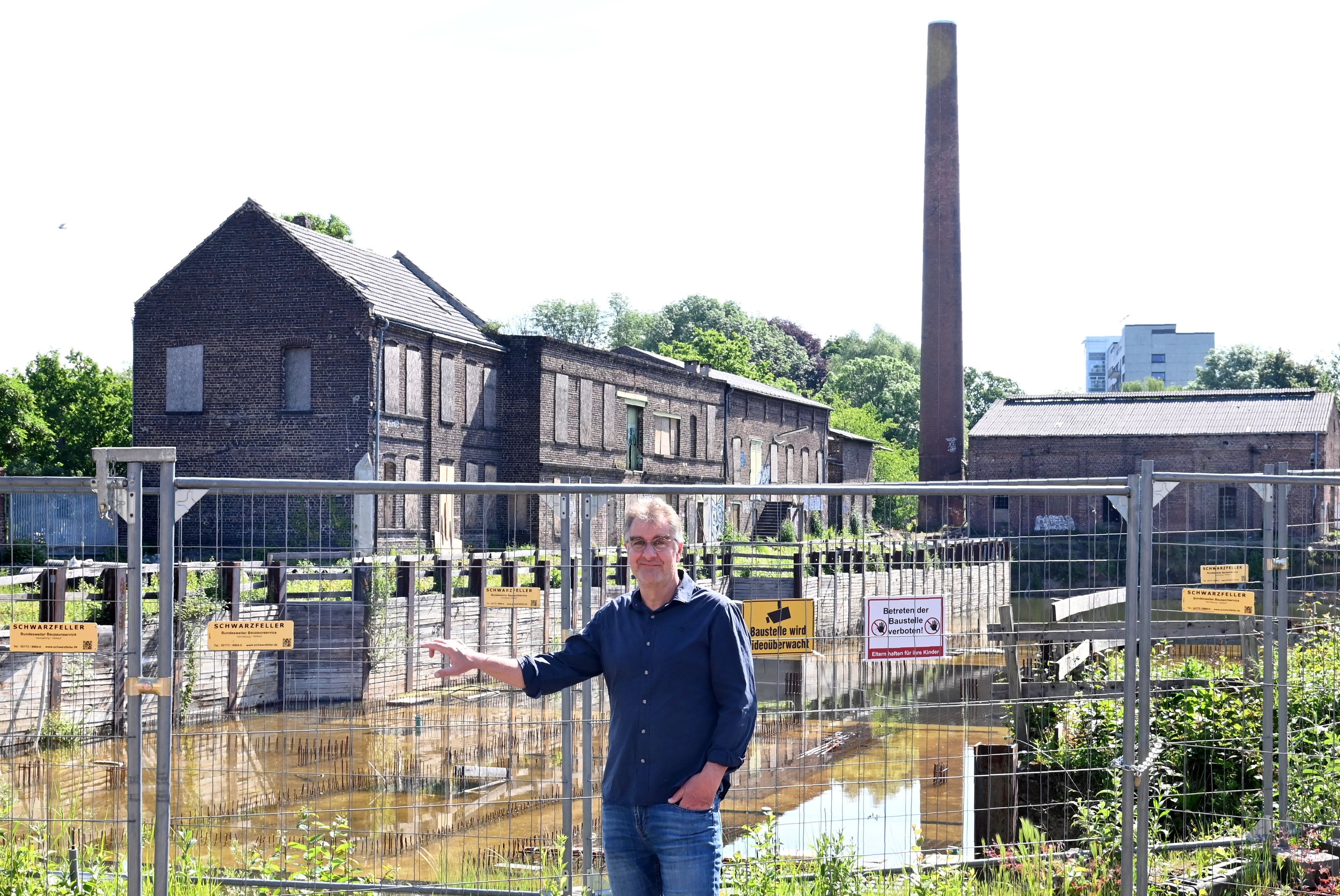 Ein Mann mit Brille, blauem Hemd und Jeans steht vor einem Bauzaun, dahinter liegen eine unfertige Tiefgarage und ein altes Backstein-Fabrikgebäude.