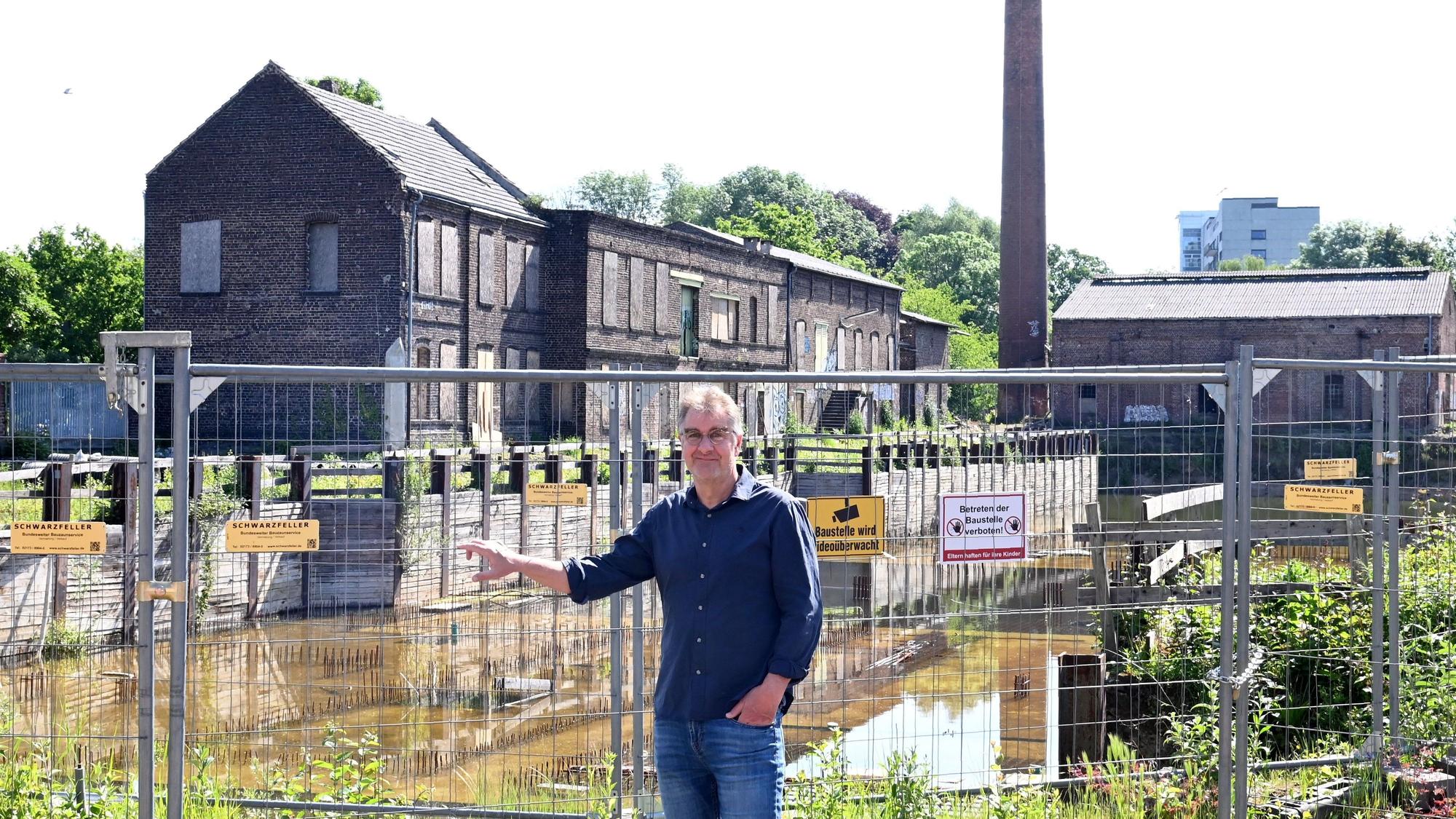 Ein Mann mit Brille, blauem Hemd und Jeans steht vor einem Bauzaun, dahinter liegen eine unfertige Tiefgarage und ein altes Backstein-Fabrikgebäude.