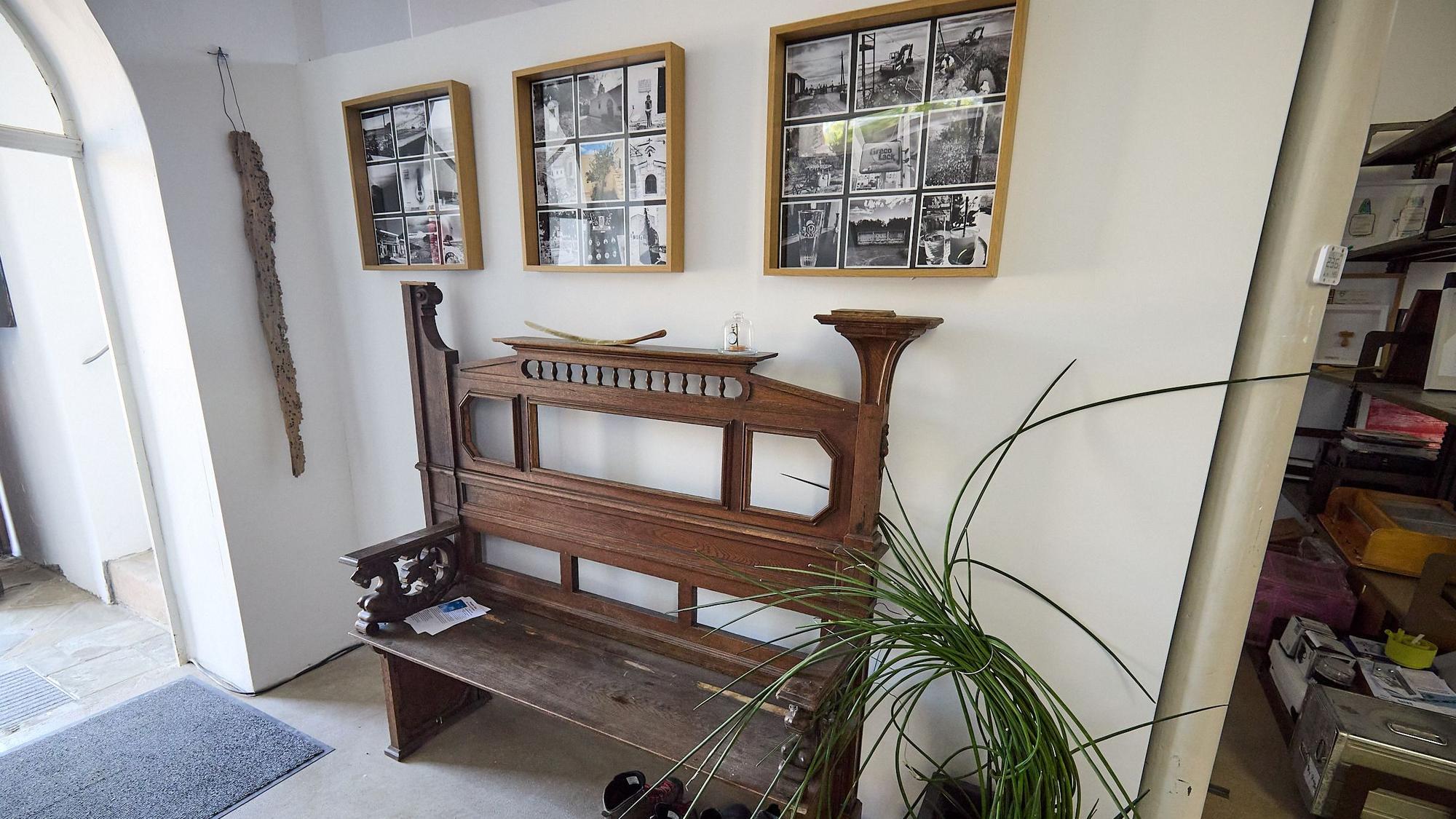An einer weiß gestrichehen Wand steht eine alte Kirchenbank, darüber hängen Fotografien,