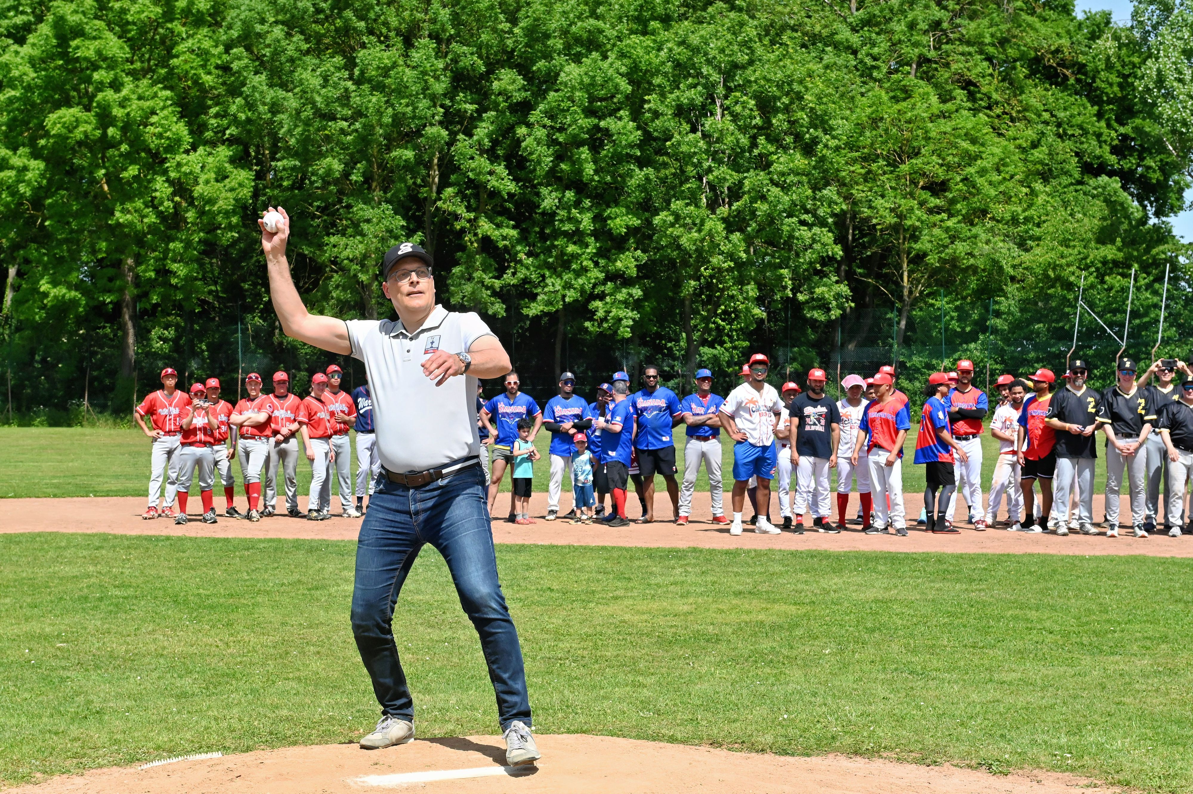 Bürgermeister Ulf Hürtgen steht auf dem Pitcherhügel und wirft einen Ball, im Hintergrund schauen mehrere Mannschaften zu.