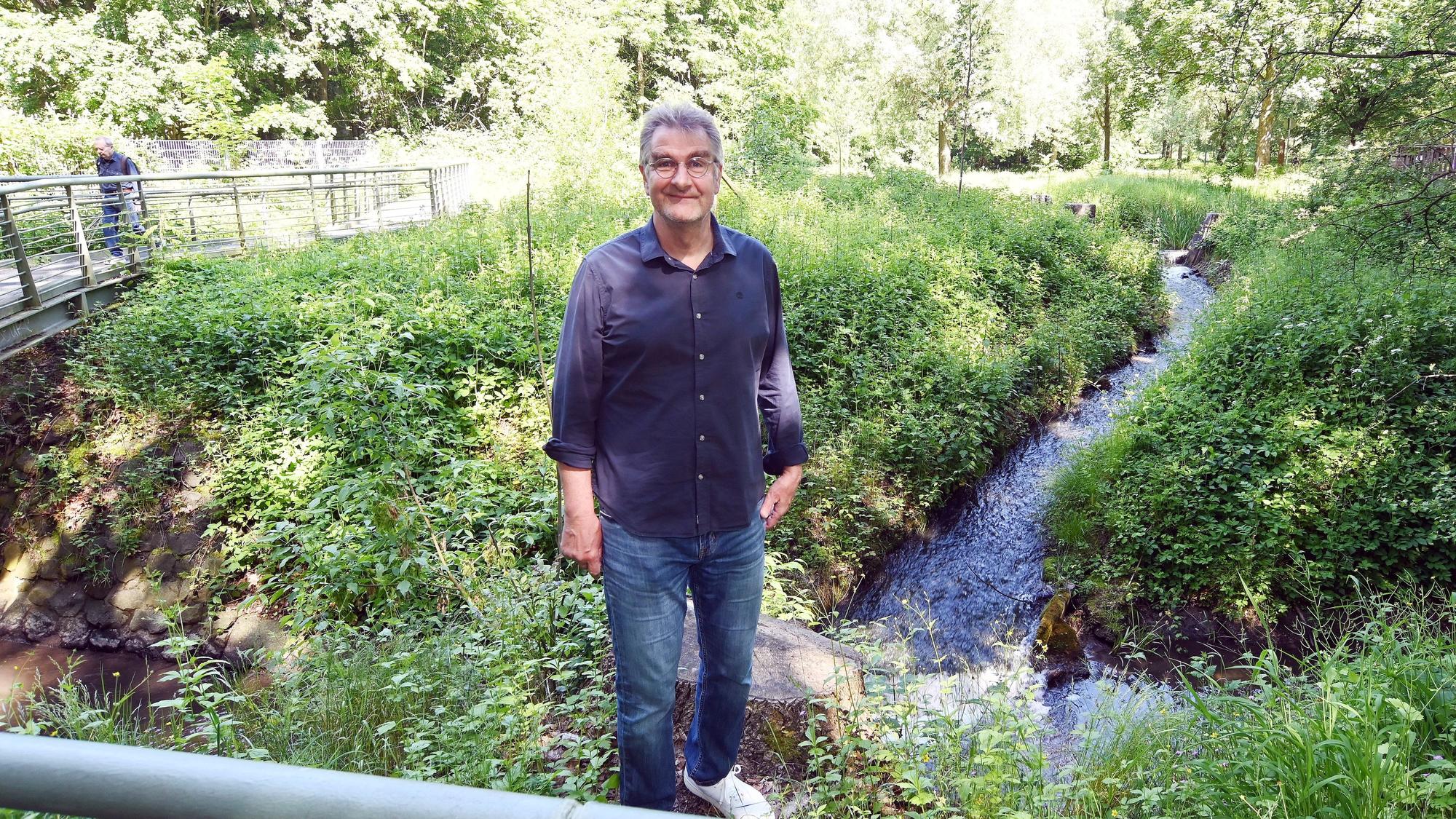 Ein Mann im blauen Hemd und Jeans steht neben einem Bachlauf.