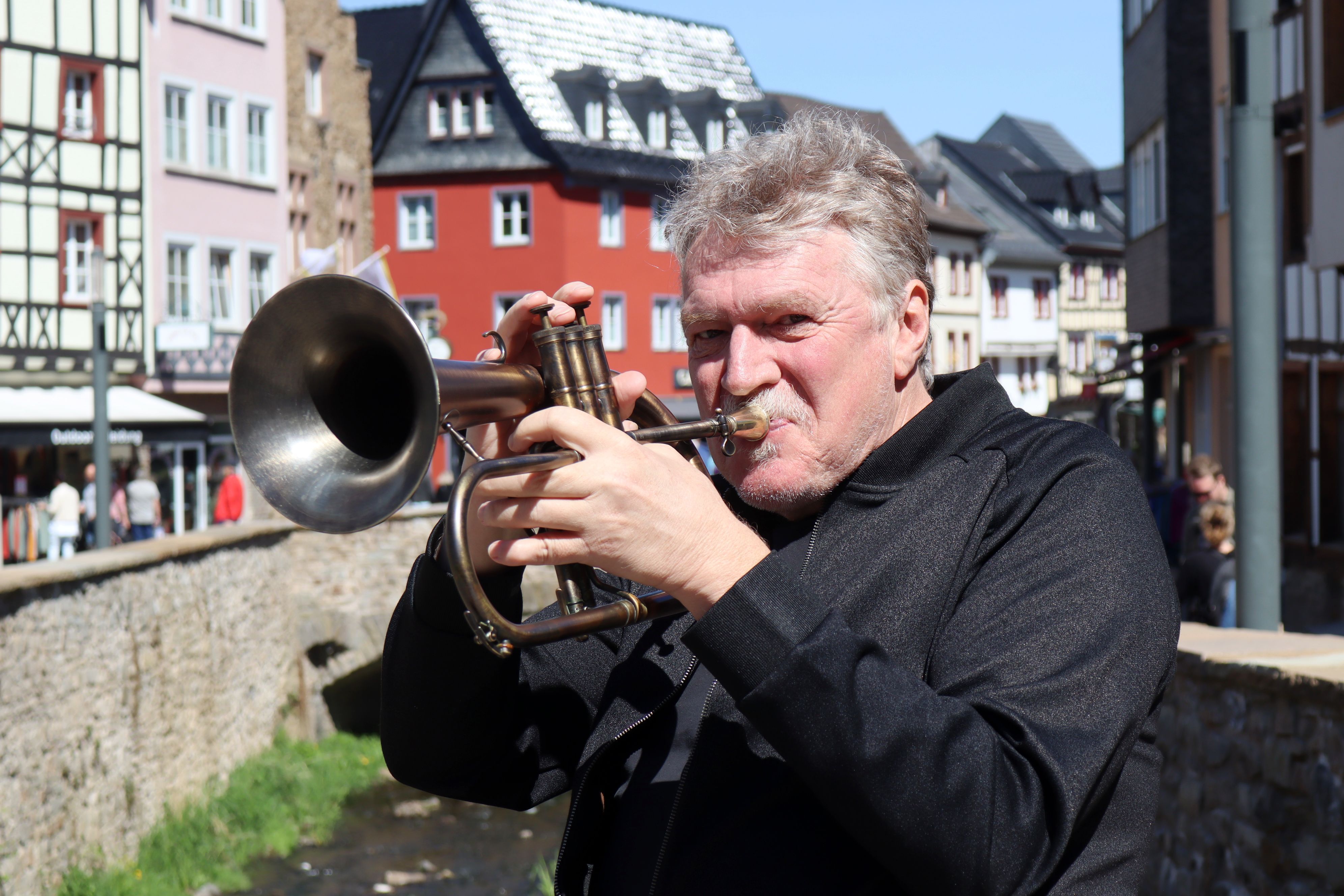 Hans Peter Salentin bläst in eine Trompete, im Hintergrund ist die Bad Münstereifeler Innenstadt zu sehen.