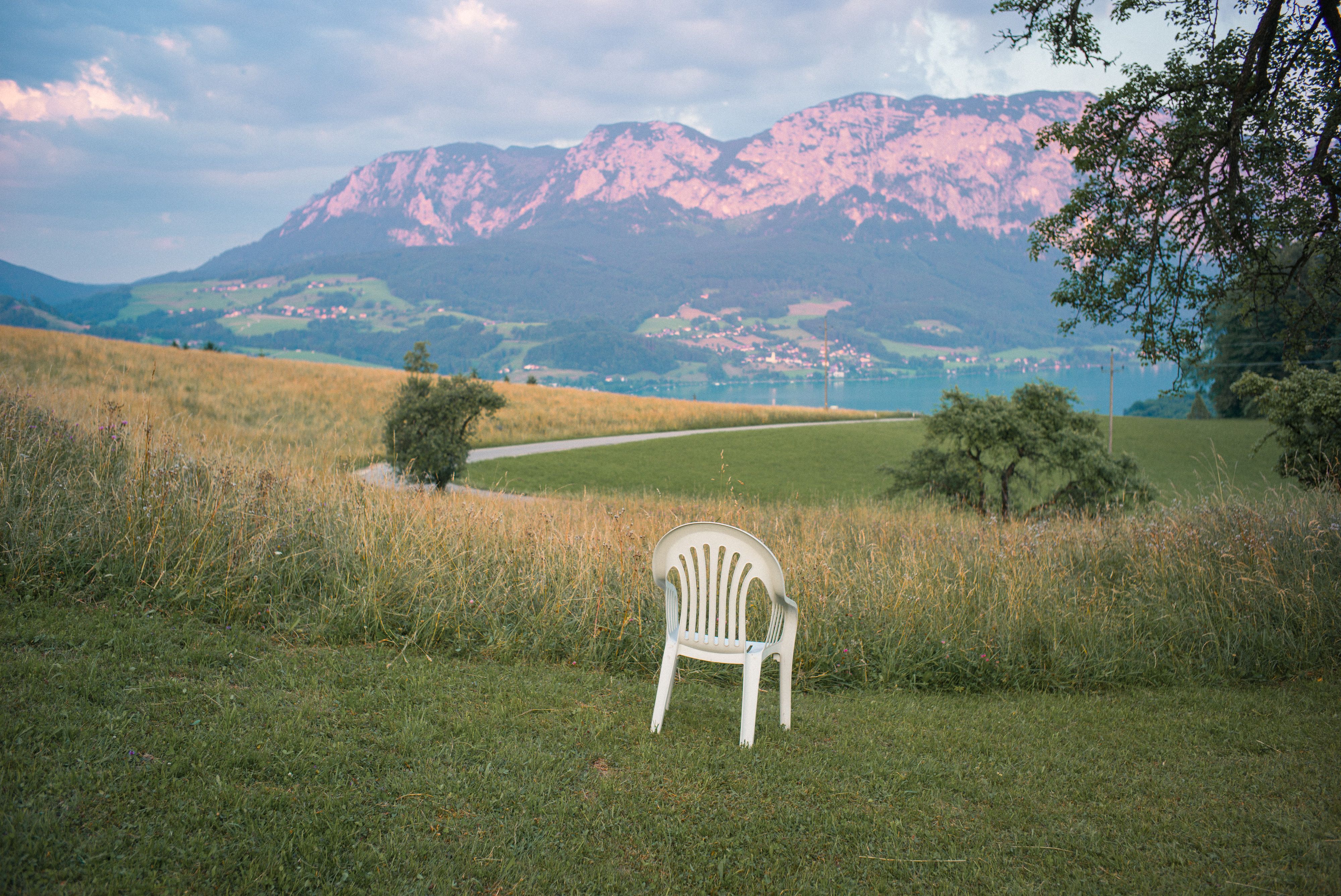 Ein weißer Monobloc-Plastikstuhl steht auf einer Wiese, im Hintergrund ist ein See vor einem Berg-Panorama zu sehen.