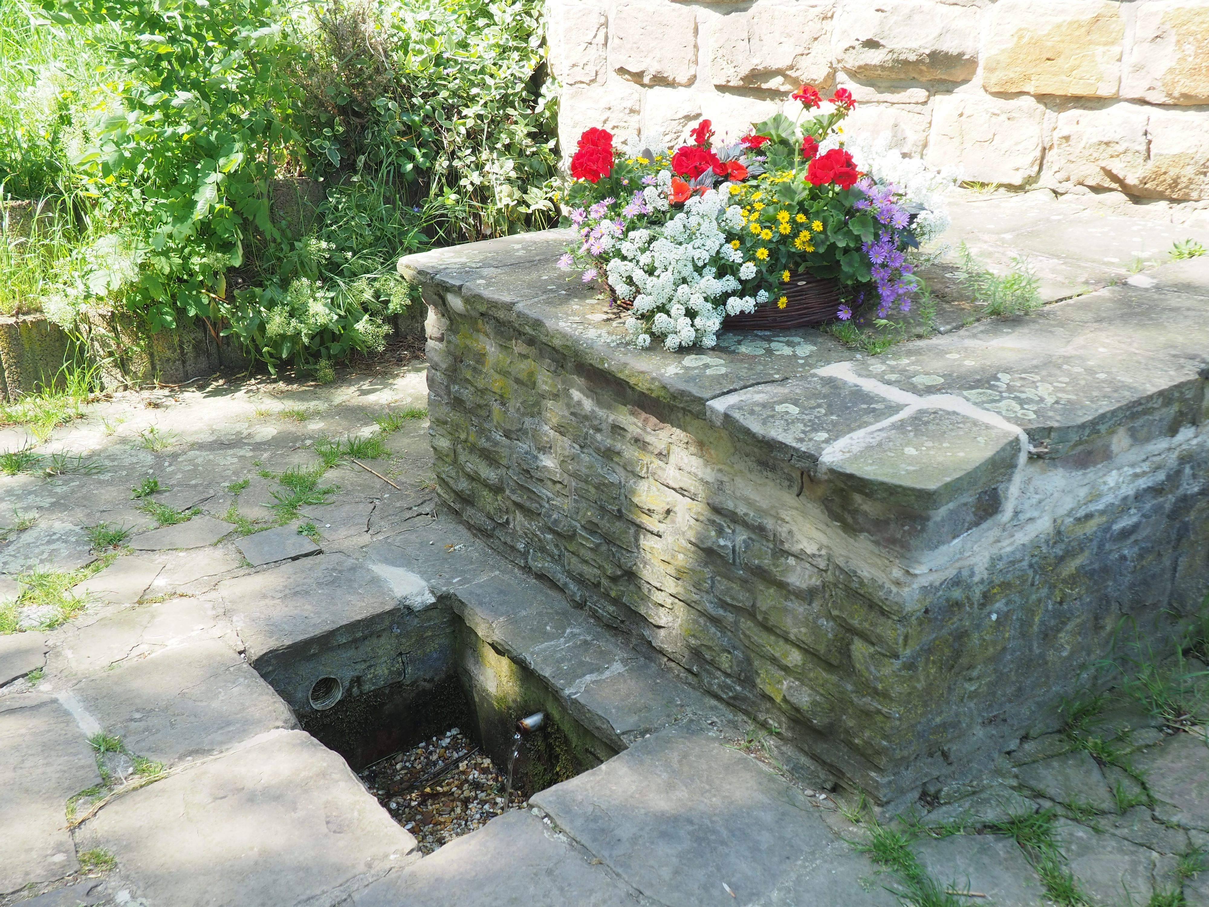 In einem kleinen, viereckigen Becken im Boden rinnt Wasser aus einem Rohr. Dahinter steht eine mit Blumen bepflanzte Schale.