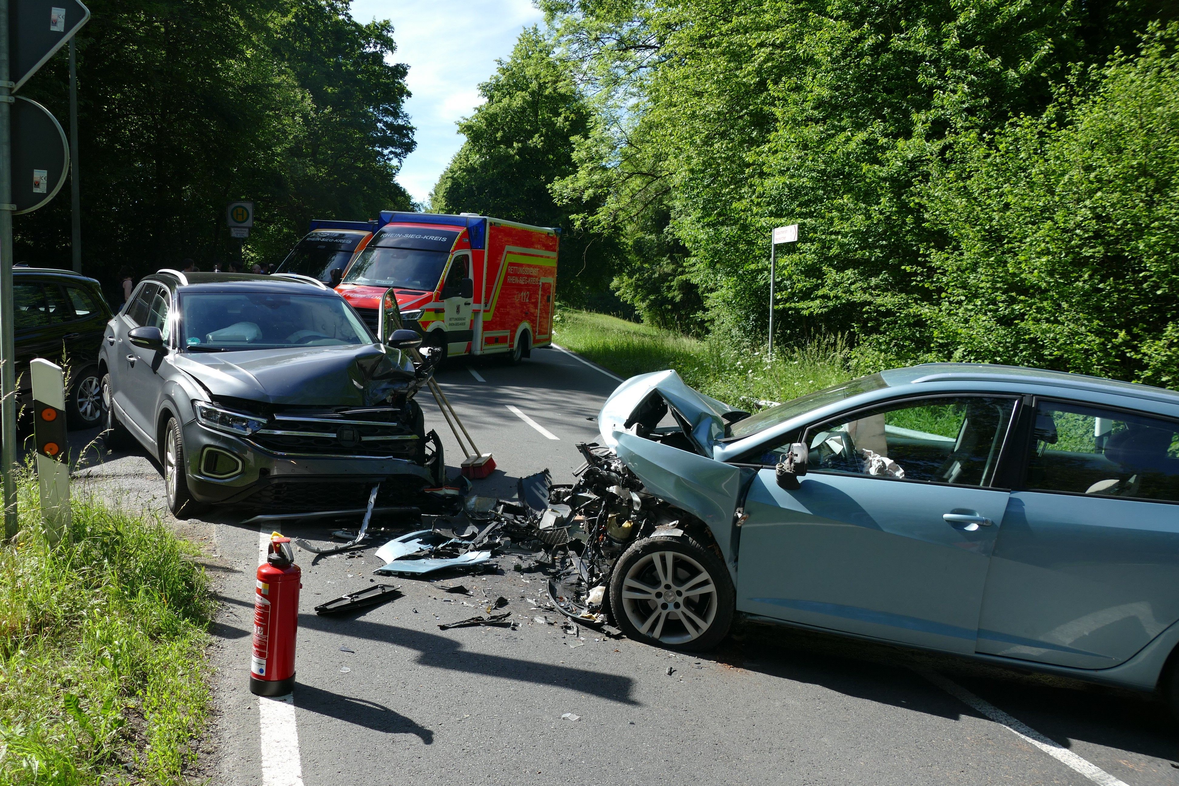 Zwei frontal zusammengestoßene Autos, zwischen ihnen Fahrzeugteile. Im Hintergrund steht ein Rettungswagen.