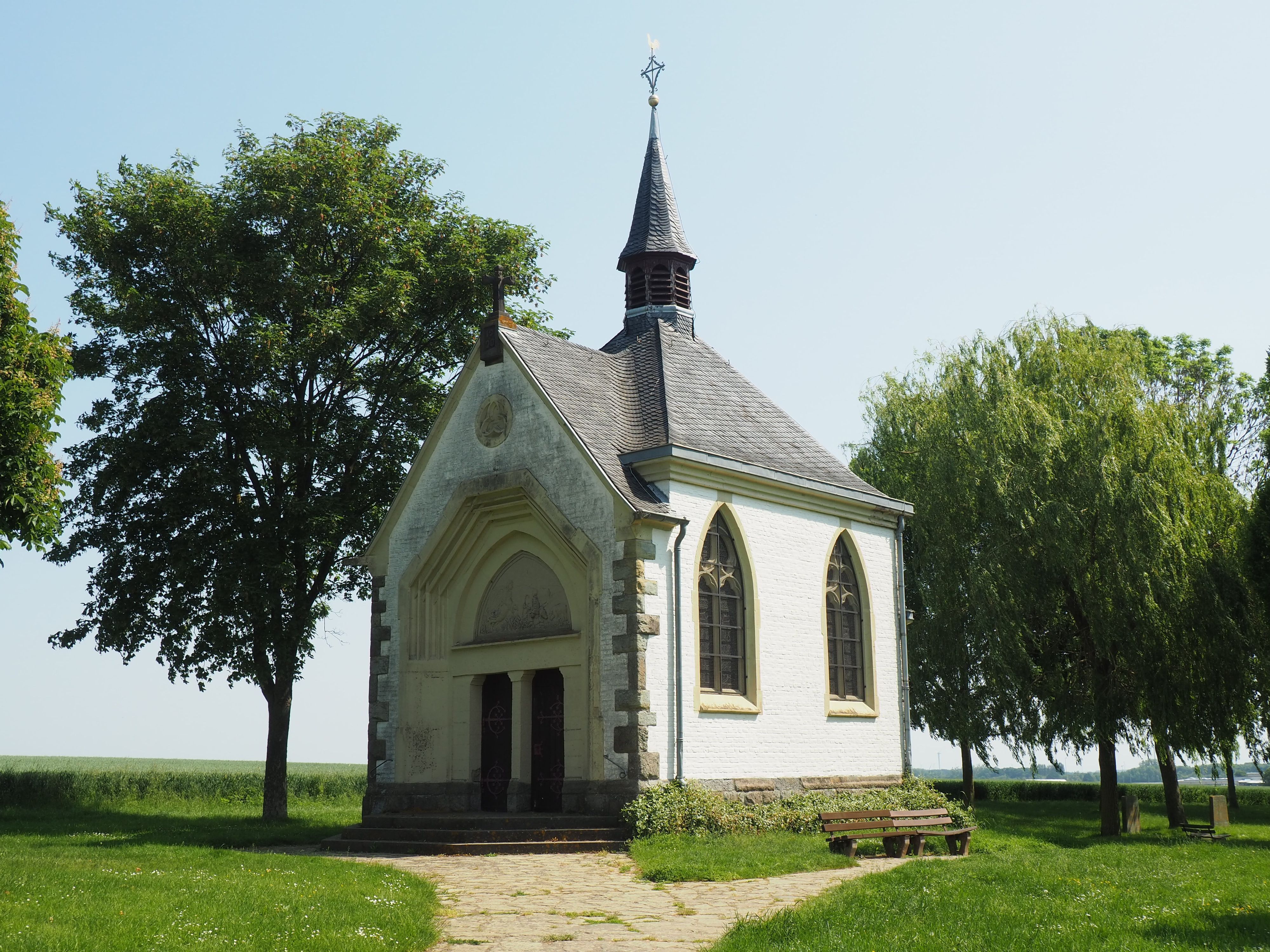 Die Kapelle steht zwischen Bäumen, ein Kiesweg führt zum Eingang.