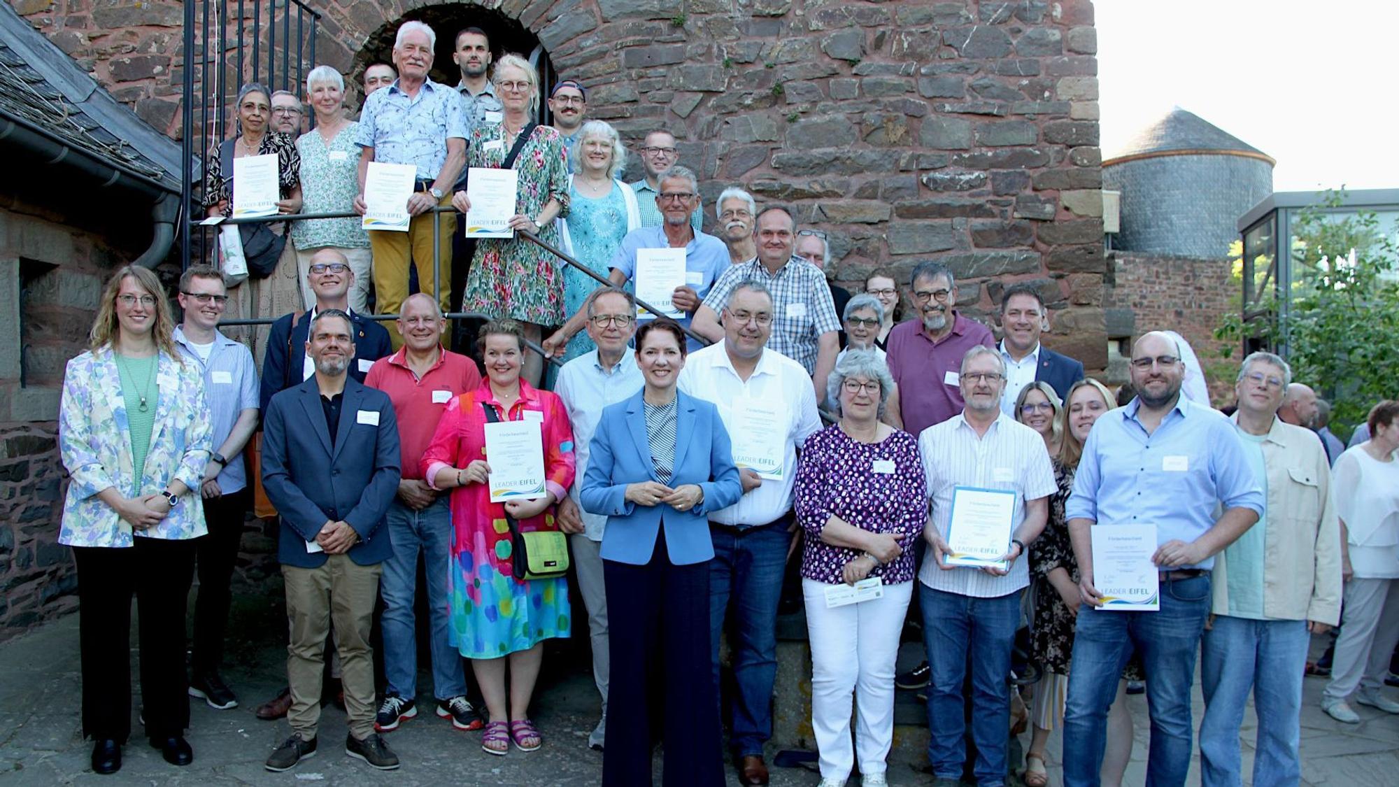 Zahlreiche Menschen stehen vor der Burg Heimbach um NRW-Ministerin, die im blauen Blazer in der ersten Reihe steht.
