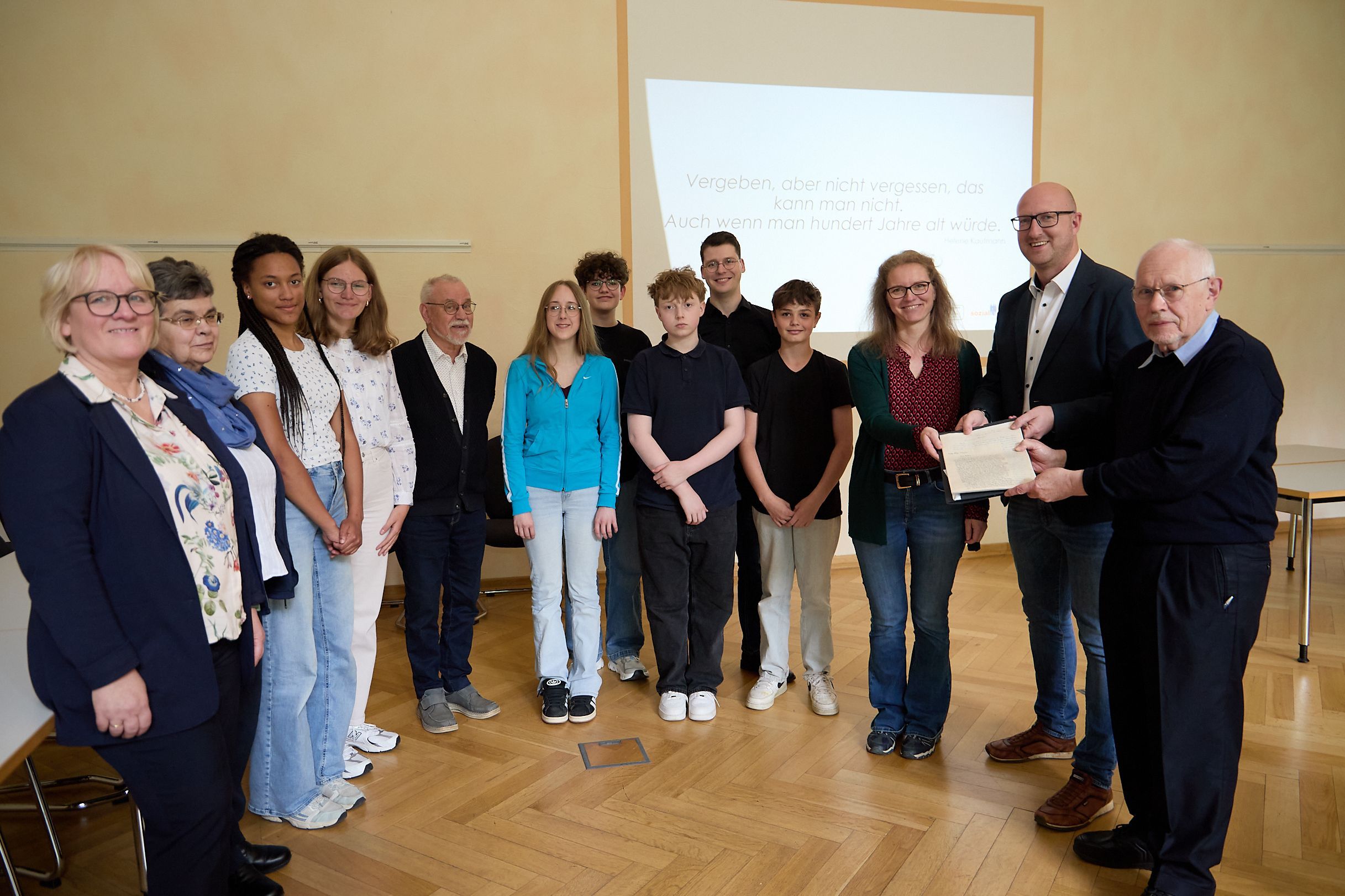 Mehrere Jugendliche und Erwachsene stehen nebeneinander. Ein Mann übergibt einem anderen und einer Frau ein Schriftstück.