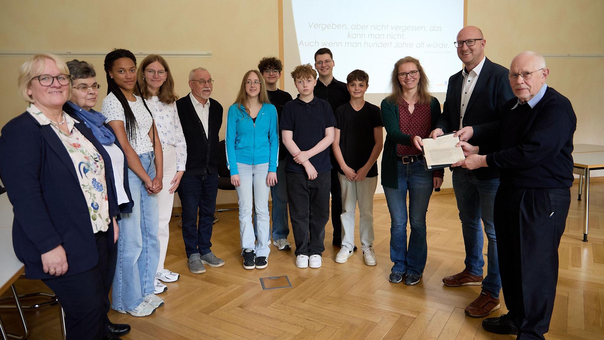 Mehrere Jugendliche und Erwachsene stehen nebeneinander. Ein Mann übergibt einem anderen und einer Frau ein Schriftstück.
