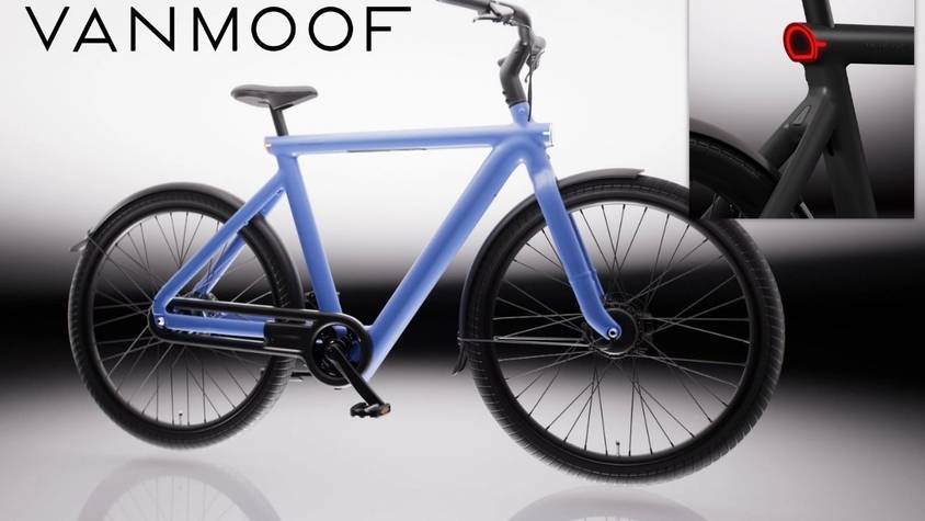 VanMoof S6 E-Bike in der Seitenansicht in der Farbe Drift Black vor hellem Hintergrund, mit Detailaufnahme des nahtlos in den Aluminiumrahmen integrierten Rücklichts in der Farbe Electric Blue – sichtbar auch das integrierte Frontlicht am Vorderrad.
