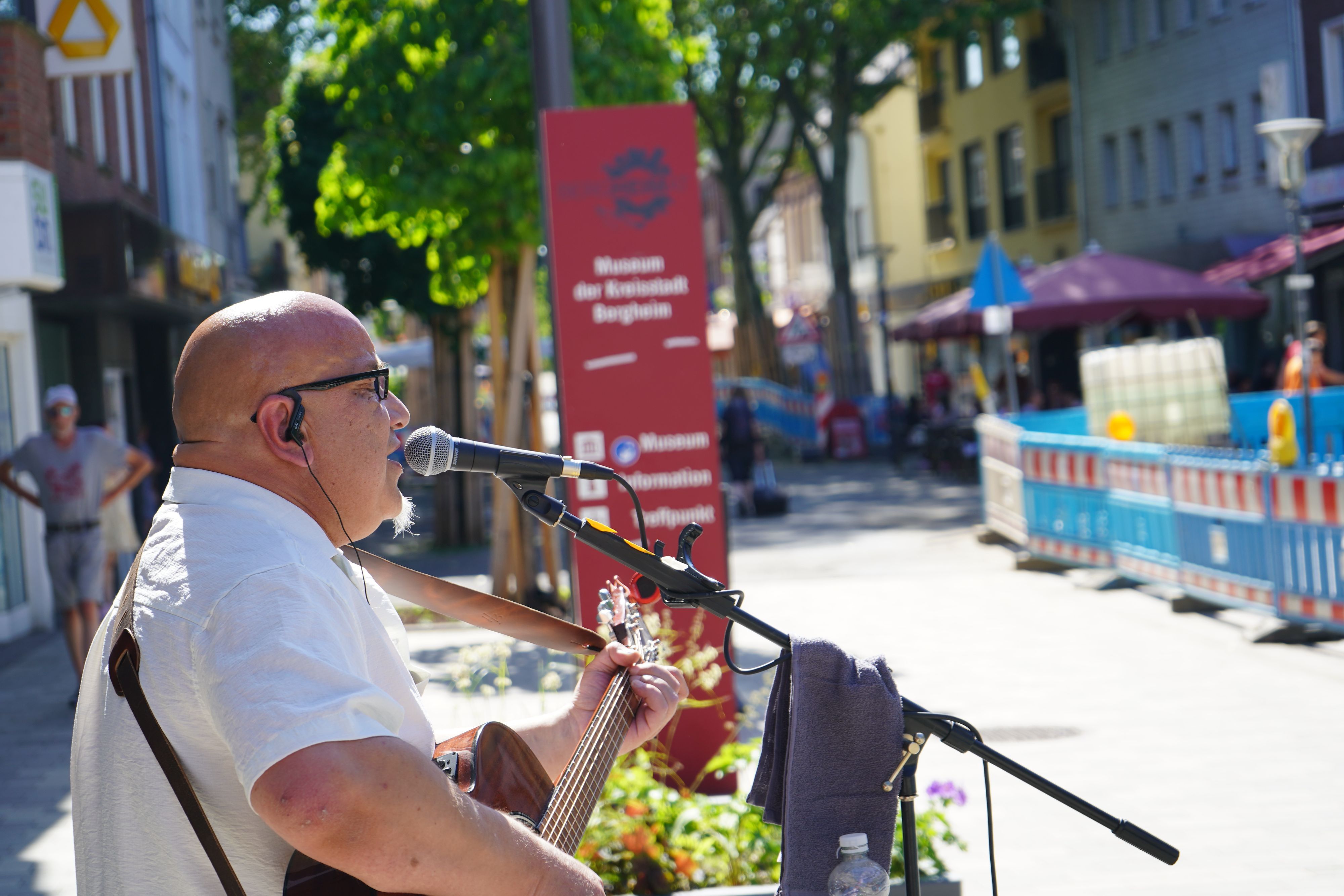 Zu sehen ist der Musiker. Er hält eine Gitarre und sinkt in ein Mikrofon. Im Hintergrund sind Bauzäune zu sehen.