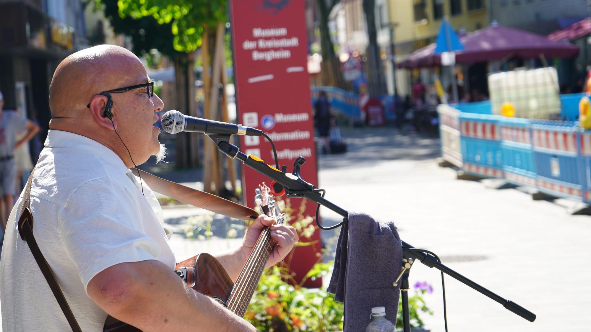 Zu sehen ist der Musiker. Er hält eine Gitarre und sinkt in ein Mikrofon. Im Hintergrund sind Bauzäune zu sehen.