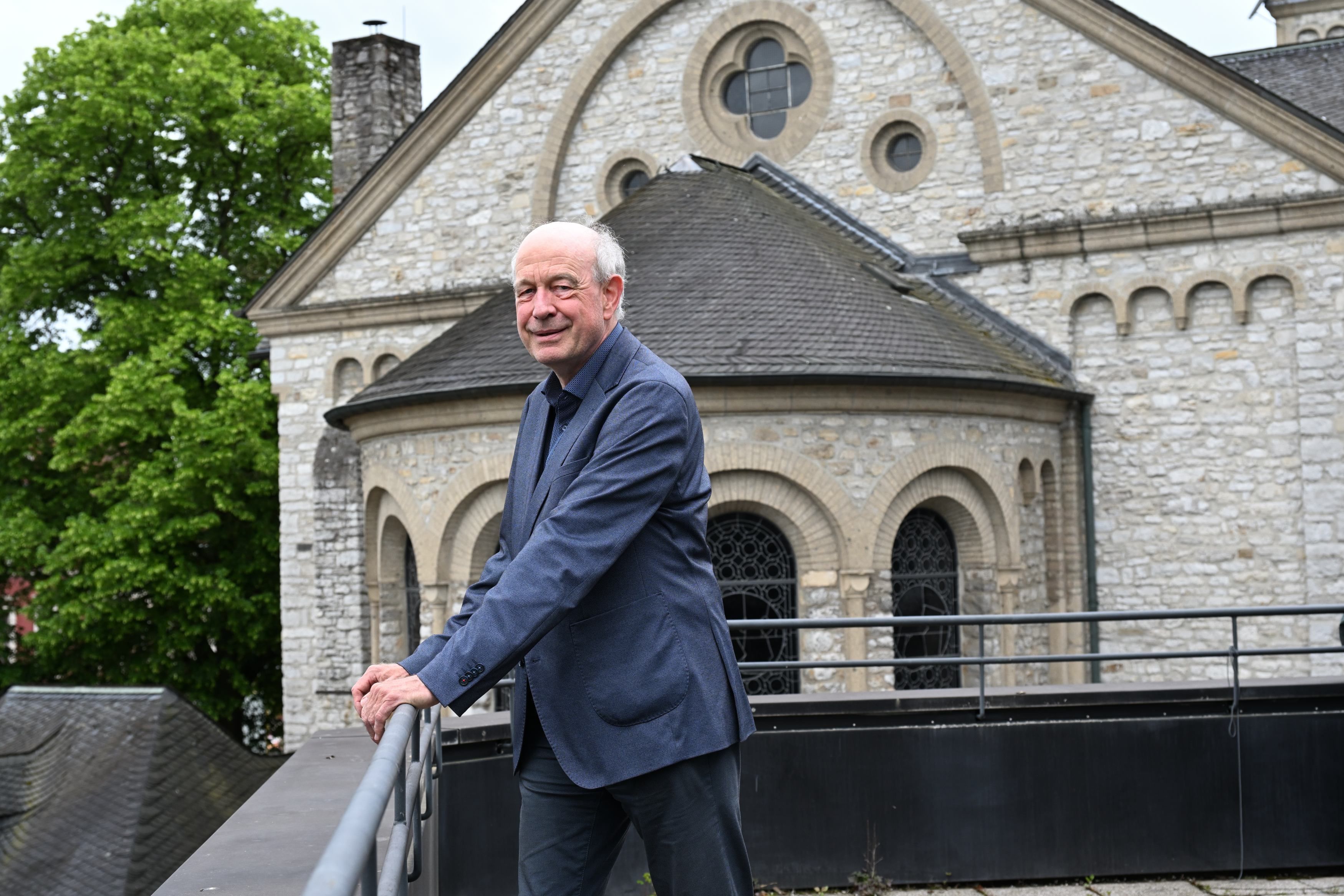 Ein Mann steht an einer Brüstung, dahinter sieht man die Apsis einer neoromanischen Kirche.