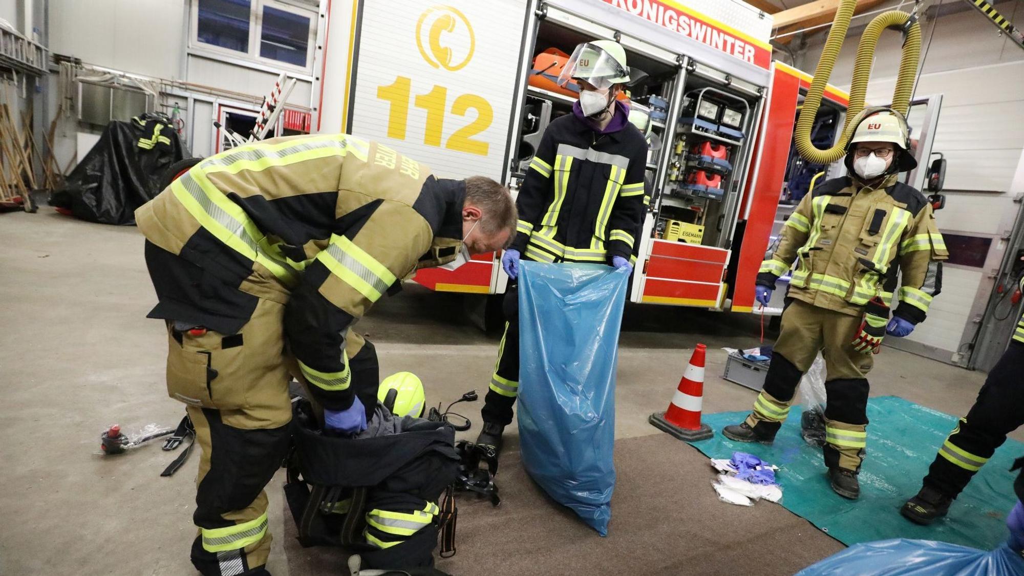 Feuerwehrleute in Schutzkleidung und mit Maske verstauen Einsatzkleidung in blauen Müllsäcken.