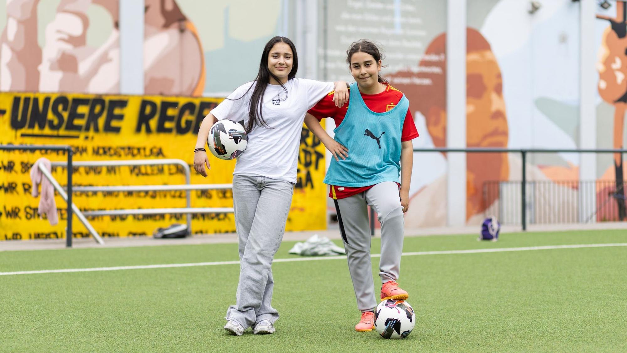 Zwei Mädchen schauen in die Kamera. Eine hat einen Ball unter ihrem Arm, die andere unter dem Fuß.