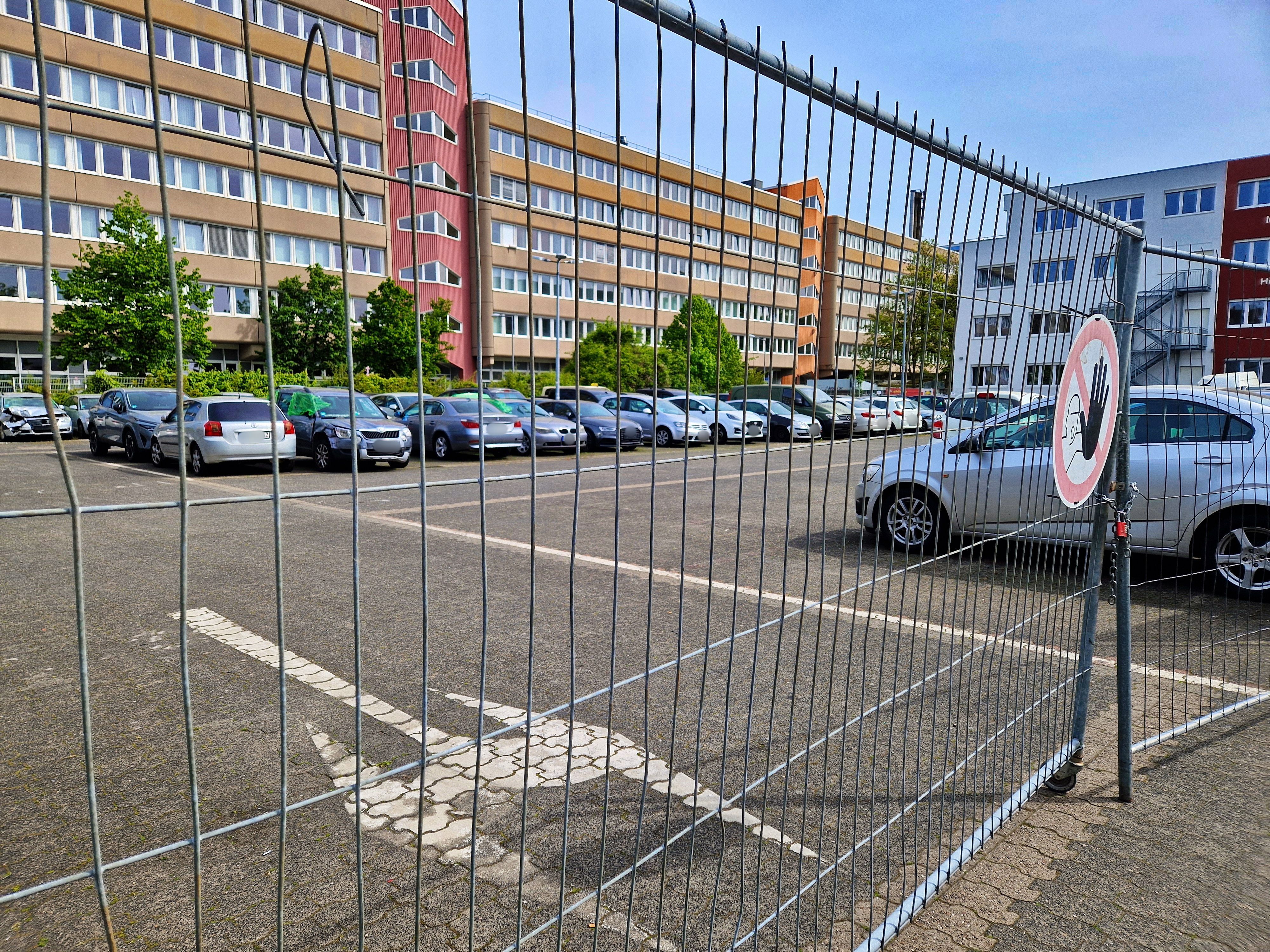 Hinter einem Bauzaun ist ein Parkplatz mit vielen abgestellten Autos.