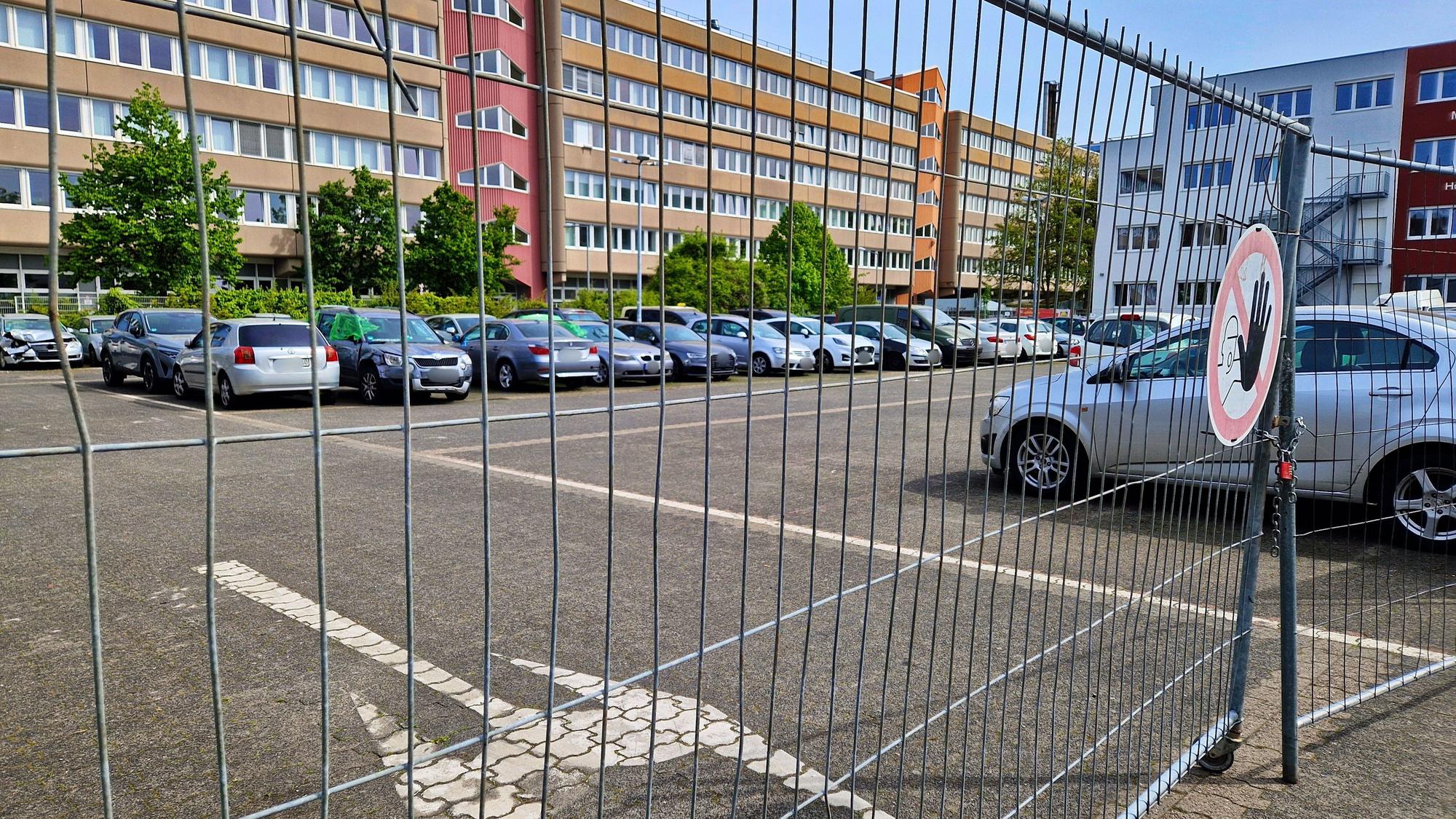 Hinter einem Bauzaun ist ein Parkplatz mit vielen abgestellten Autos.