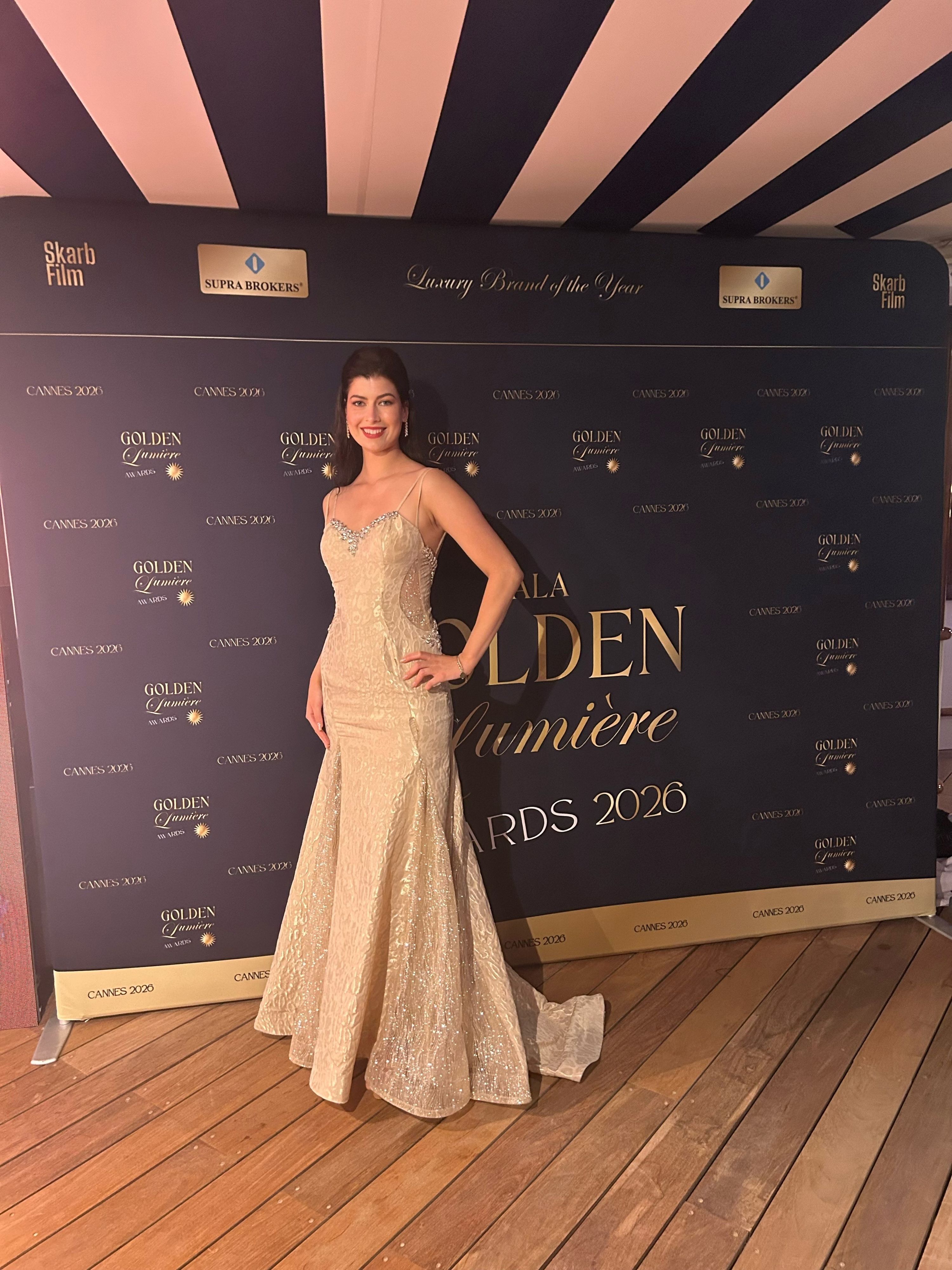 Eine Frau im goldenen Abendkleid steht vor einer dunklen Wand mit goldener Schrift.