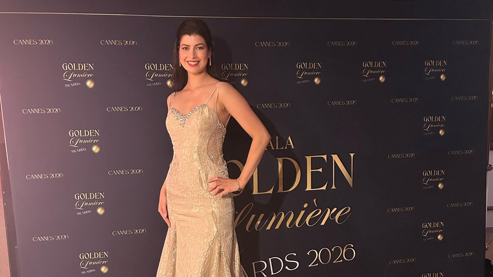 Eine Frau im goldenen Abendkleid steht vor einer dunklen Wand mit goldener Schrift.