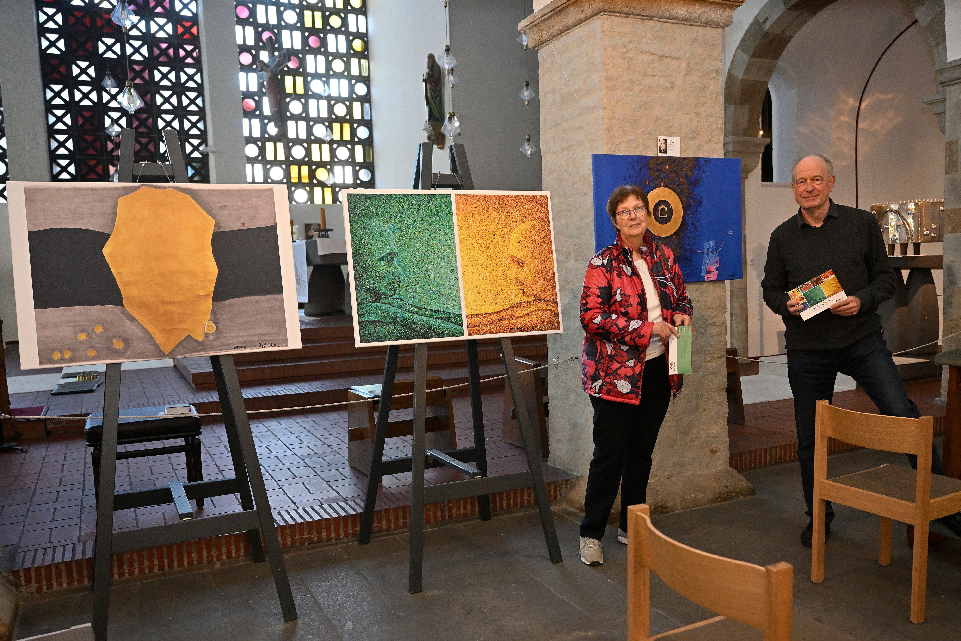 Bilder von Hunger- oder Fastentüchern werden in einer Kirche ausgestellt. Daneben stehen ein Mann und eine Frau.