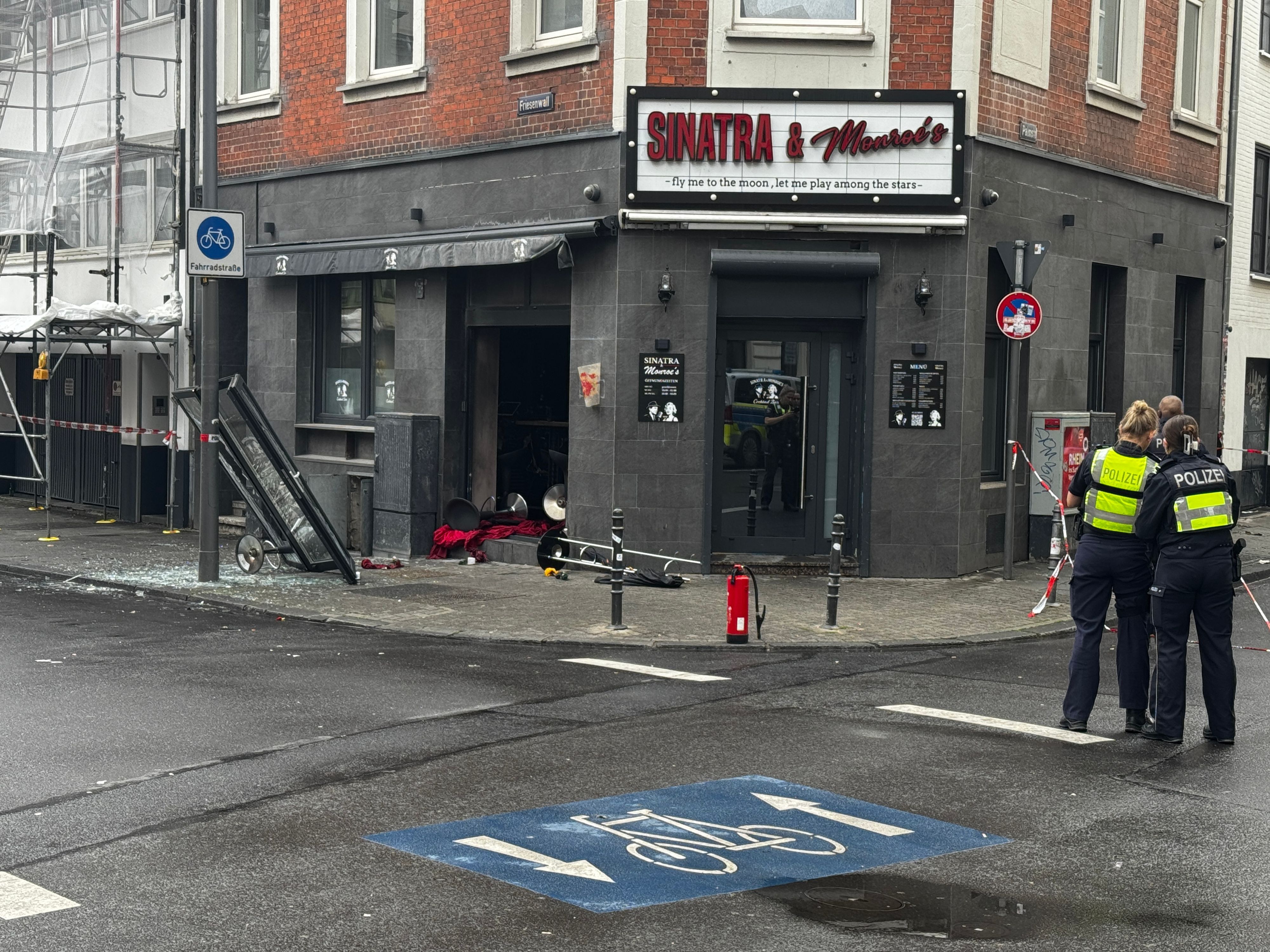 Blick auf die beschädigte Bar mit zerstörter Scheibe, Trümmern auf dem Boden und einem Feuerlöscher davor. Im Vordergrund stehen zwei Polizeibeamte mit Warnwesten an einem abgesperrten Bereich.