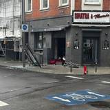Blick auf die beschädigte Bar mit zerstörter Scheibe, Trümmern auf dem Boden und einem Feuerlöscher davor. Im Vordergrund stehen zwei Polizeibeamte mit Warnwesten an einem abgesperrten Bereich.