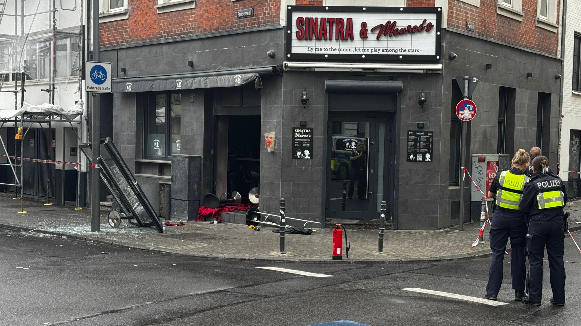 Blick auf die beschädigte Bar "Sinatra & Monroe's" mit eingeschlagener Scheibe, Trümmern auf dem Boden und einem Feuerlöscher davor. Im Vordergrund stehen zwei Polizeibeamte mit Warnwesten an einem abgesperrten Bereich. Friesenwall, Köln